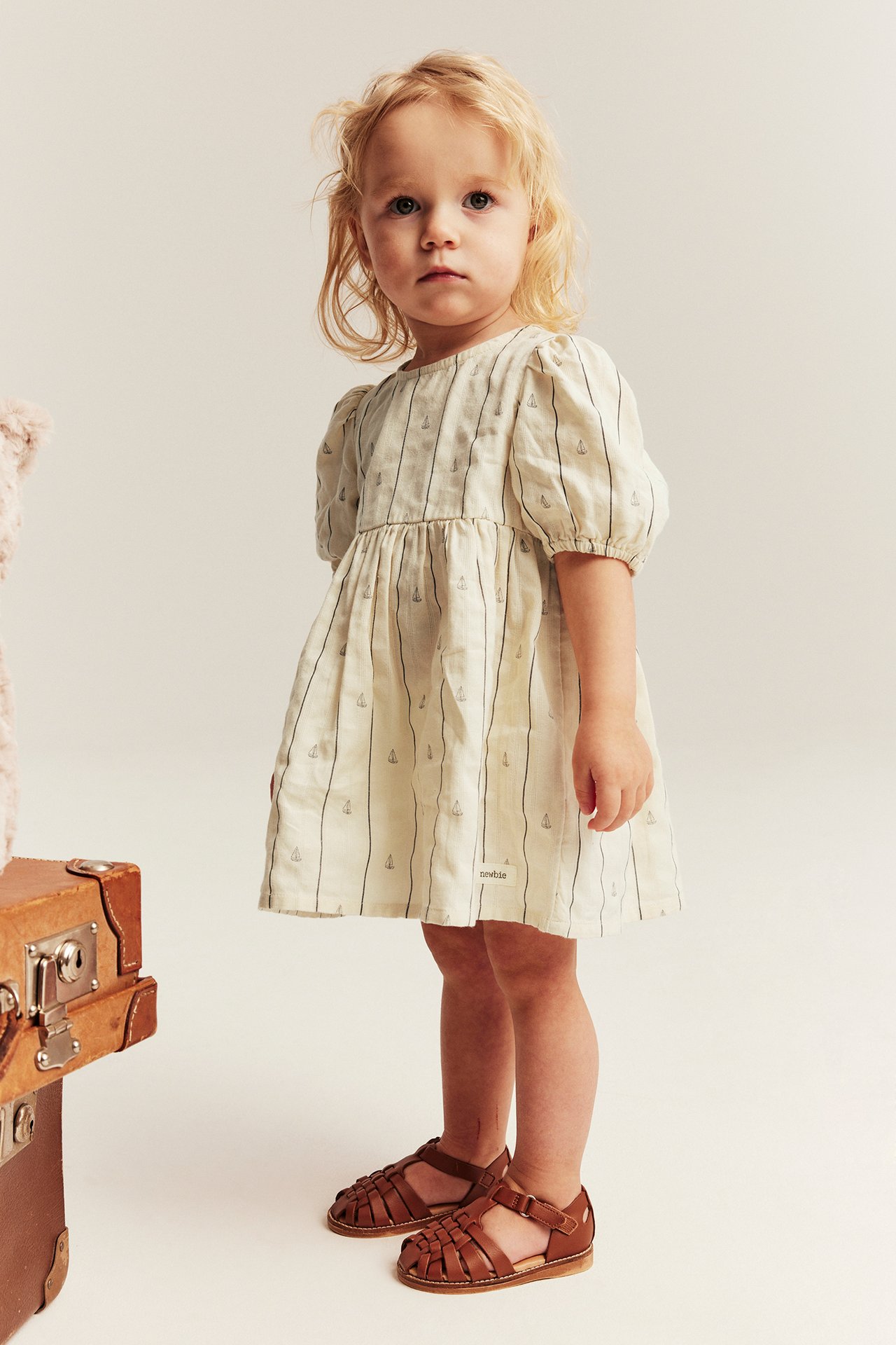 Edestä: Vauvatyttö valkoisessa raidallisessa puuvillamekossa purjeveneillä, puffihihoilla ja ruskeilla sandaaleilla.