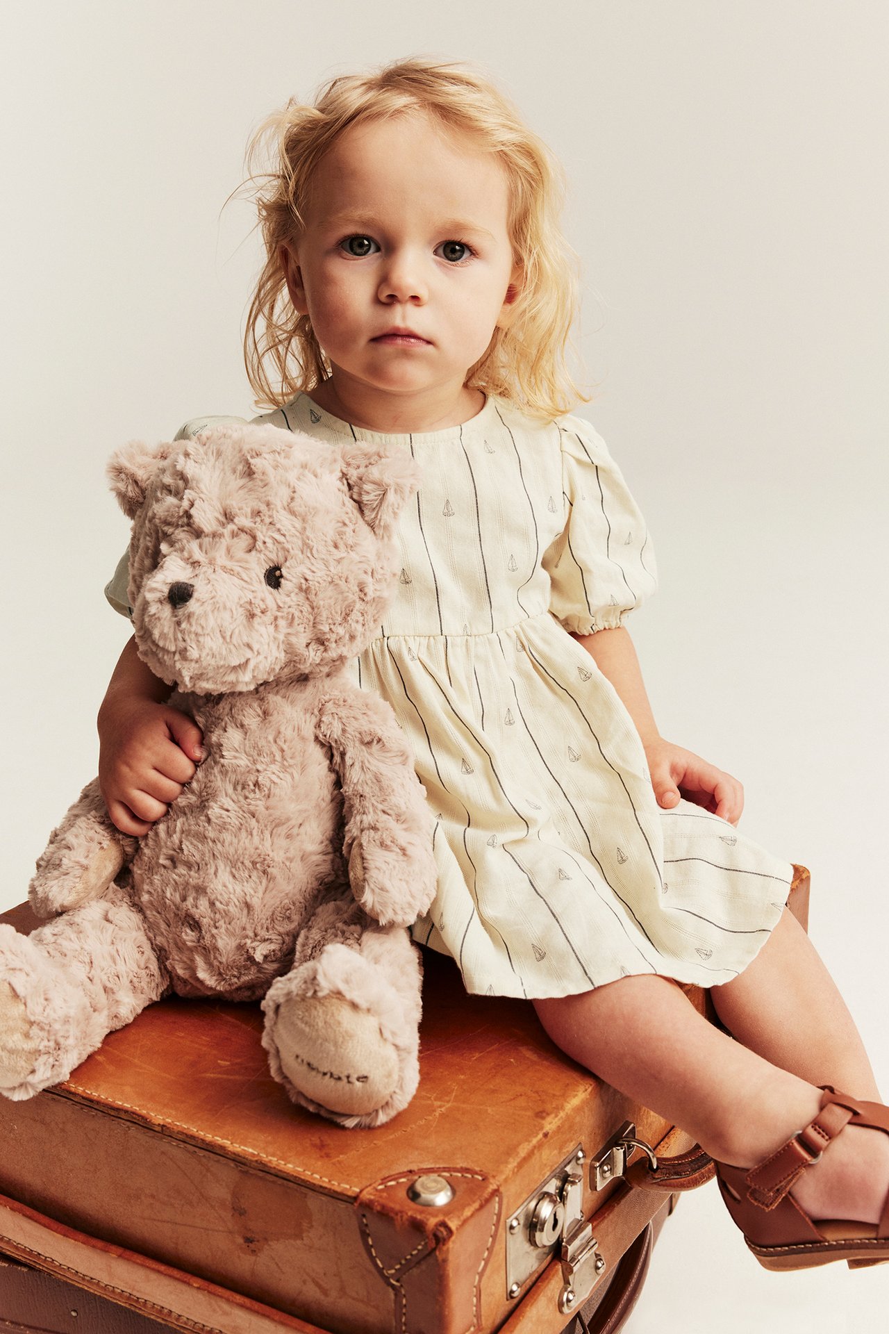 Vauva valkoisessa raidallisessa venekuvioisessa mekossa, puhvihihat, ruskeat sandaalit, pitää nallea.