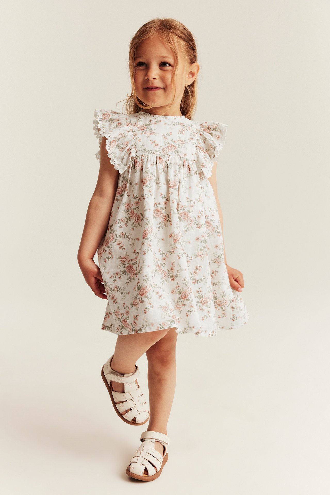 Forfra, et barn i en offwhite blomstret volangkjole med blondekanter og offwhite sandaler.
