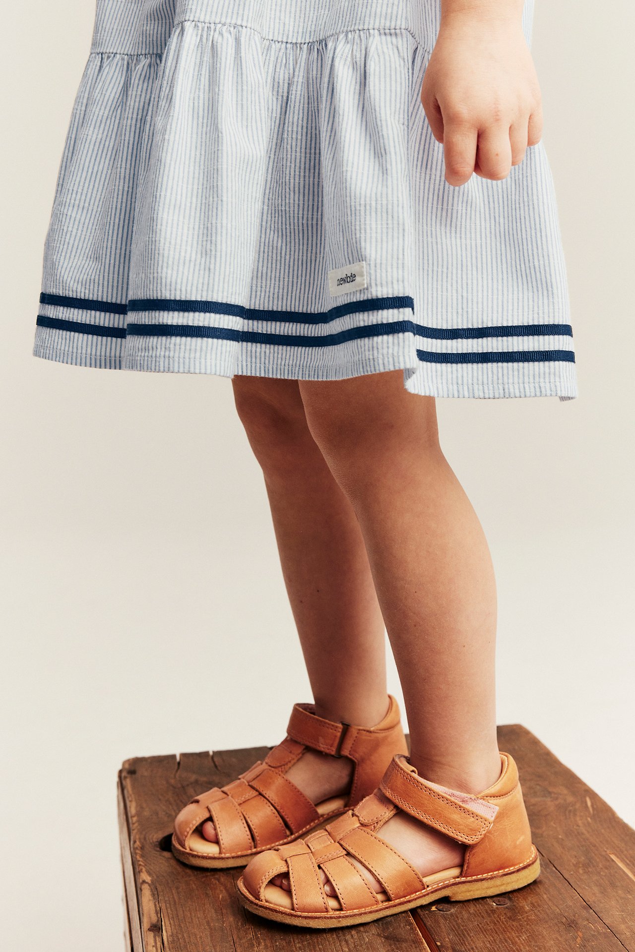 Framsiden: Barn i blå- og hvitstripete bomullskjole med marineblå bånd og brune skinnandaler.