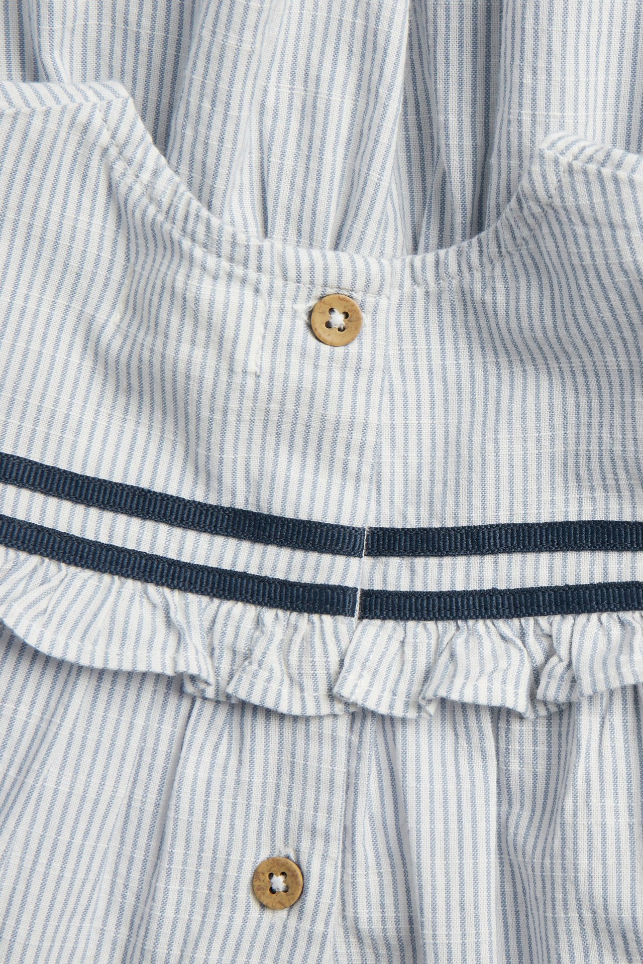 Nærbilde av barnekjole i blå-hvitstripet bomull med volang, to marineblå bånd og treknapper.
