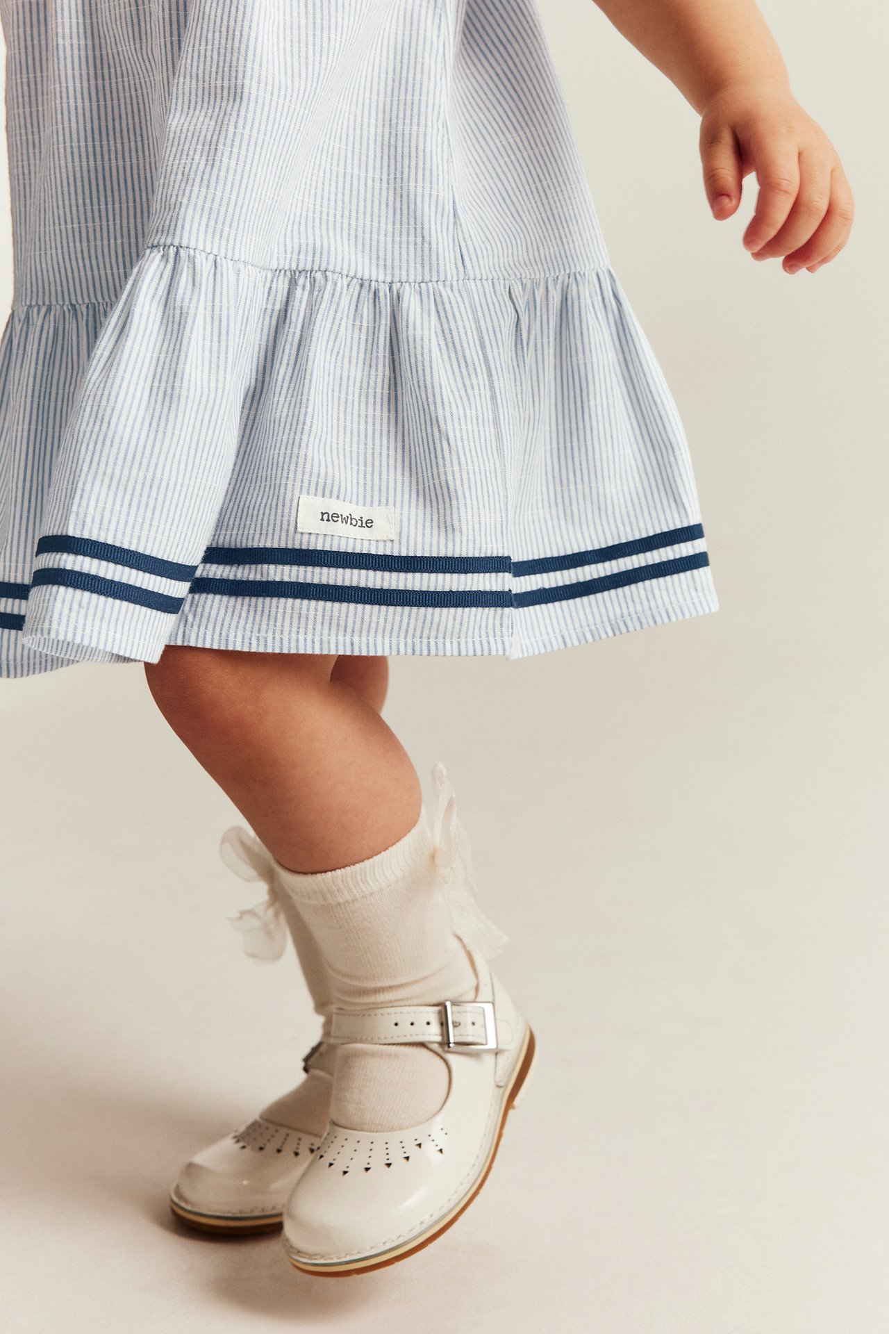 Częściowy widok z przodu: dziecko w niebieskiej sukience w paski, granatowe wykończenie, kremowe skarpetki, białe buciki Mary Jane.