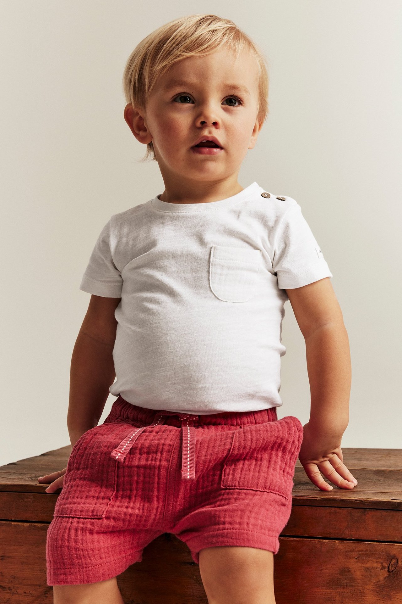 Widok z przodu: Dziecko w kremowym body z zatrzaskami na ramieniu, kieszonką i czerwonych szortach.