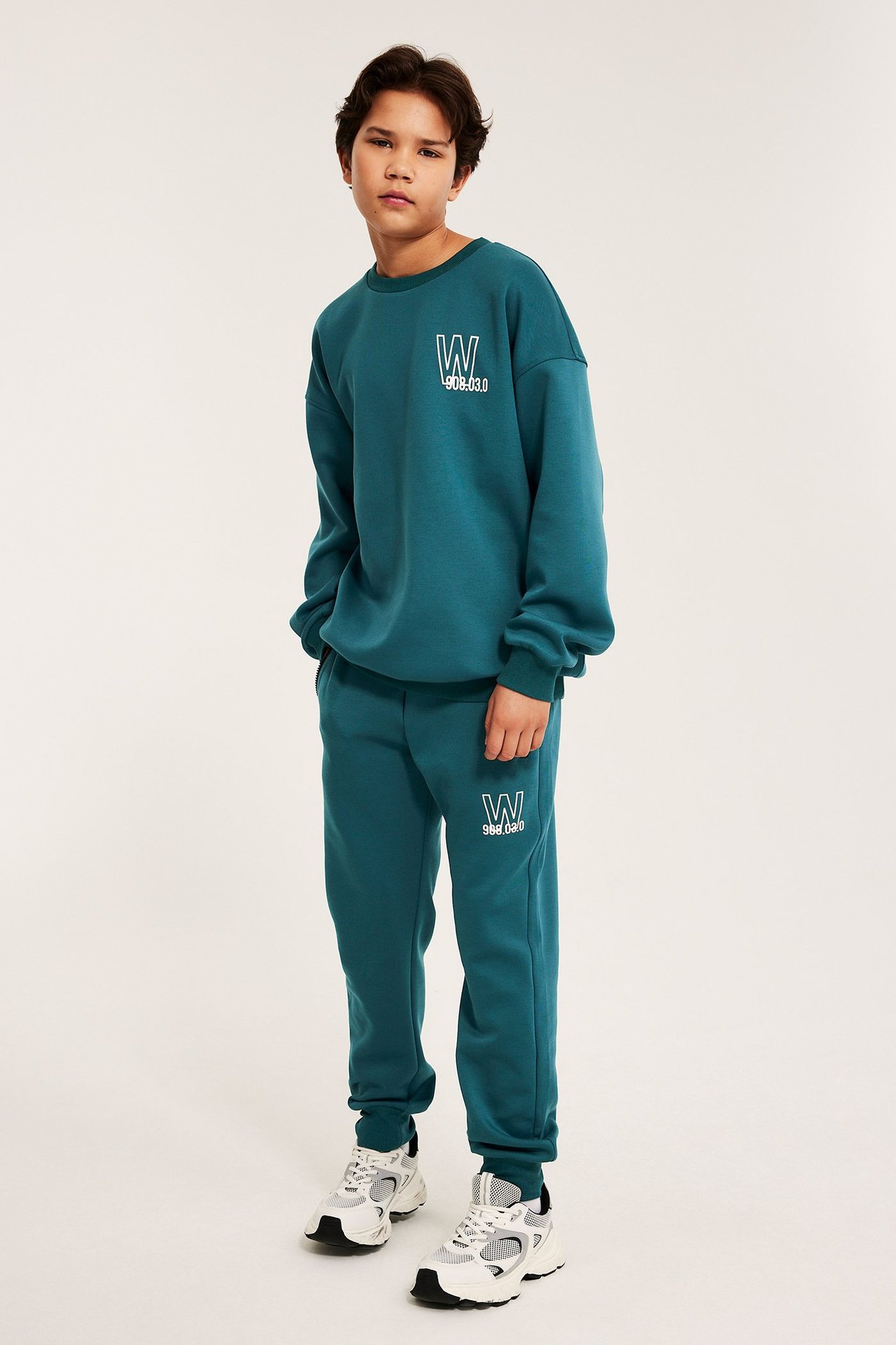 Framifrån: Ett barn i grön sweatshirt med vitt tryck, matchande sweatpants och vita sneakers.