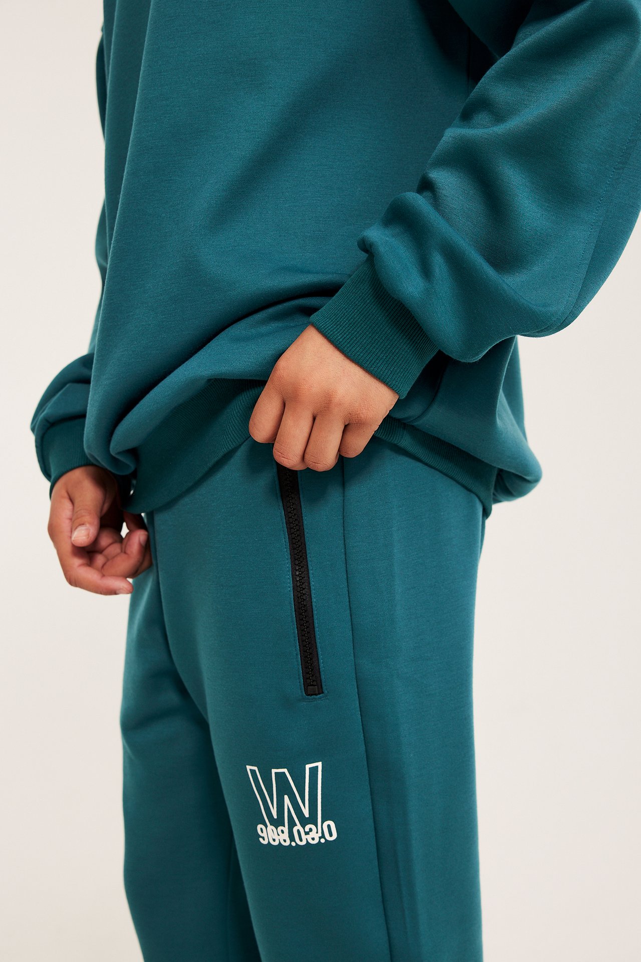 Nærbilde sidevisning: barn i grønn genser og matchende joggebukse med svart sidelås og hvitt W-trykk.