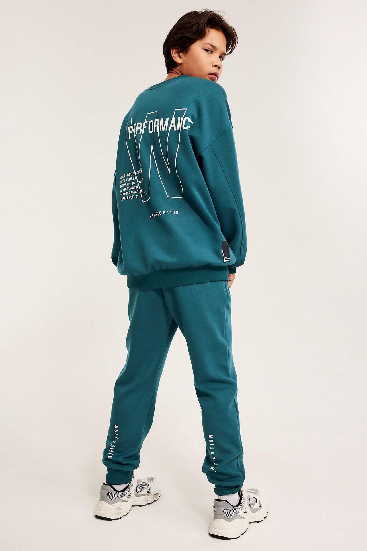 Bakifrån-vänster vy av ett barn i grönt joggingställ med vita tryck, samt sneakers.