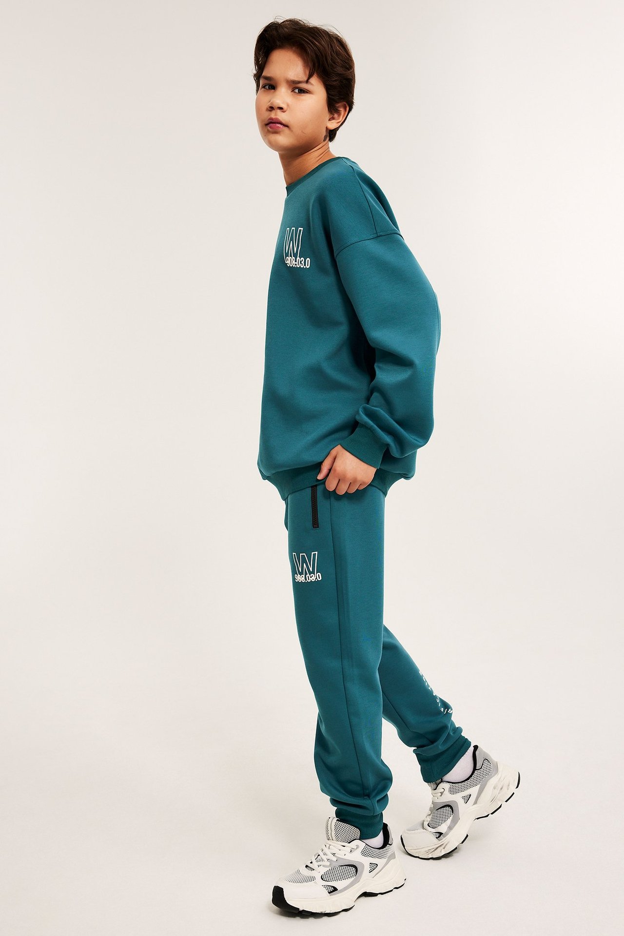 Etusivukuva lapsesta petrolinvihreässä scubamateriaalisessa verryttelyasussa, valkoisilla painatuksilla ja valkoisilla tennareilla.