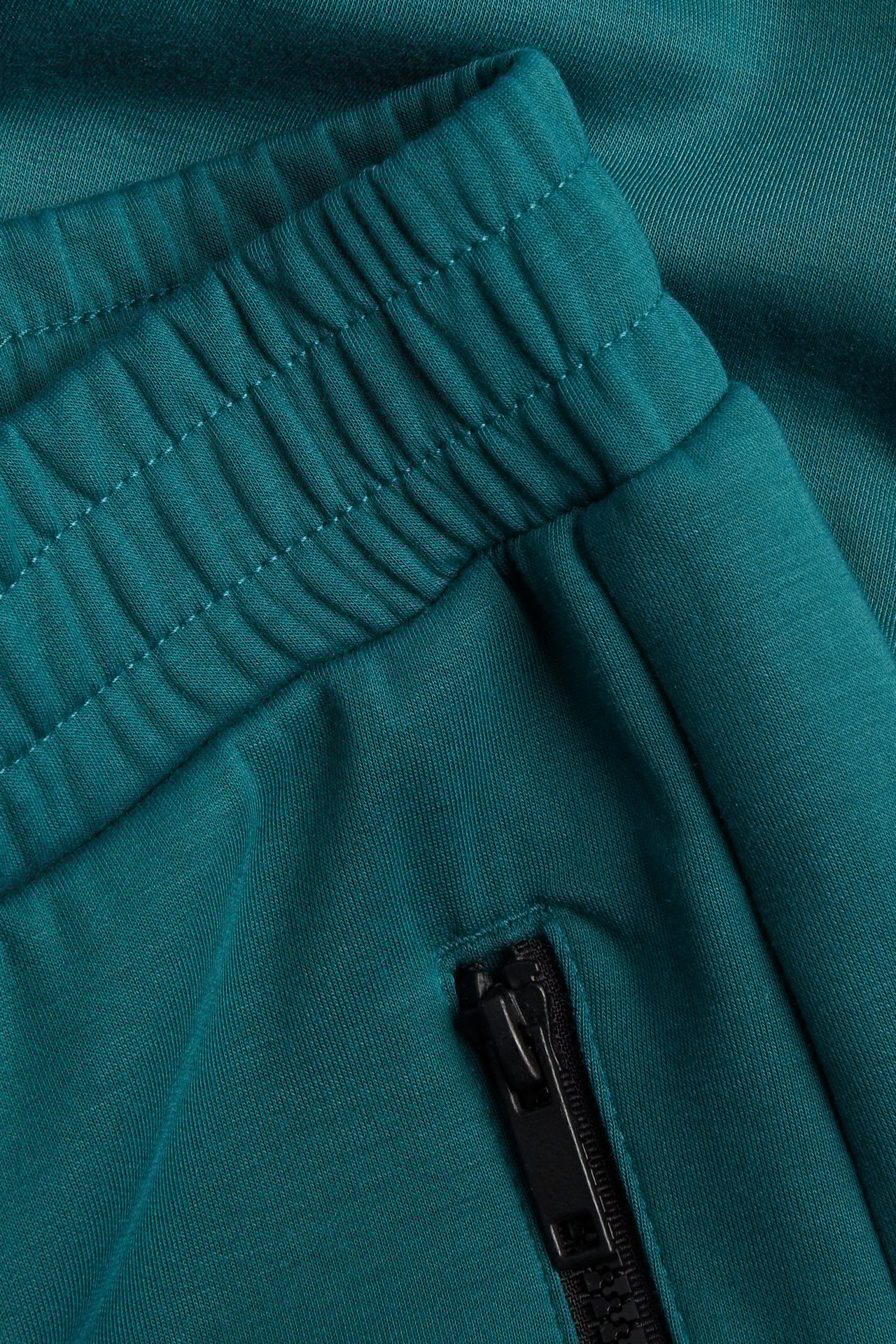 Zbliżenie zielonych spodni dresowych scuba z elastycznym pasem i czarną kieszenią na suwak.