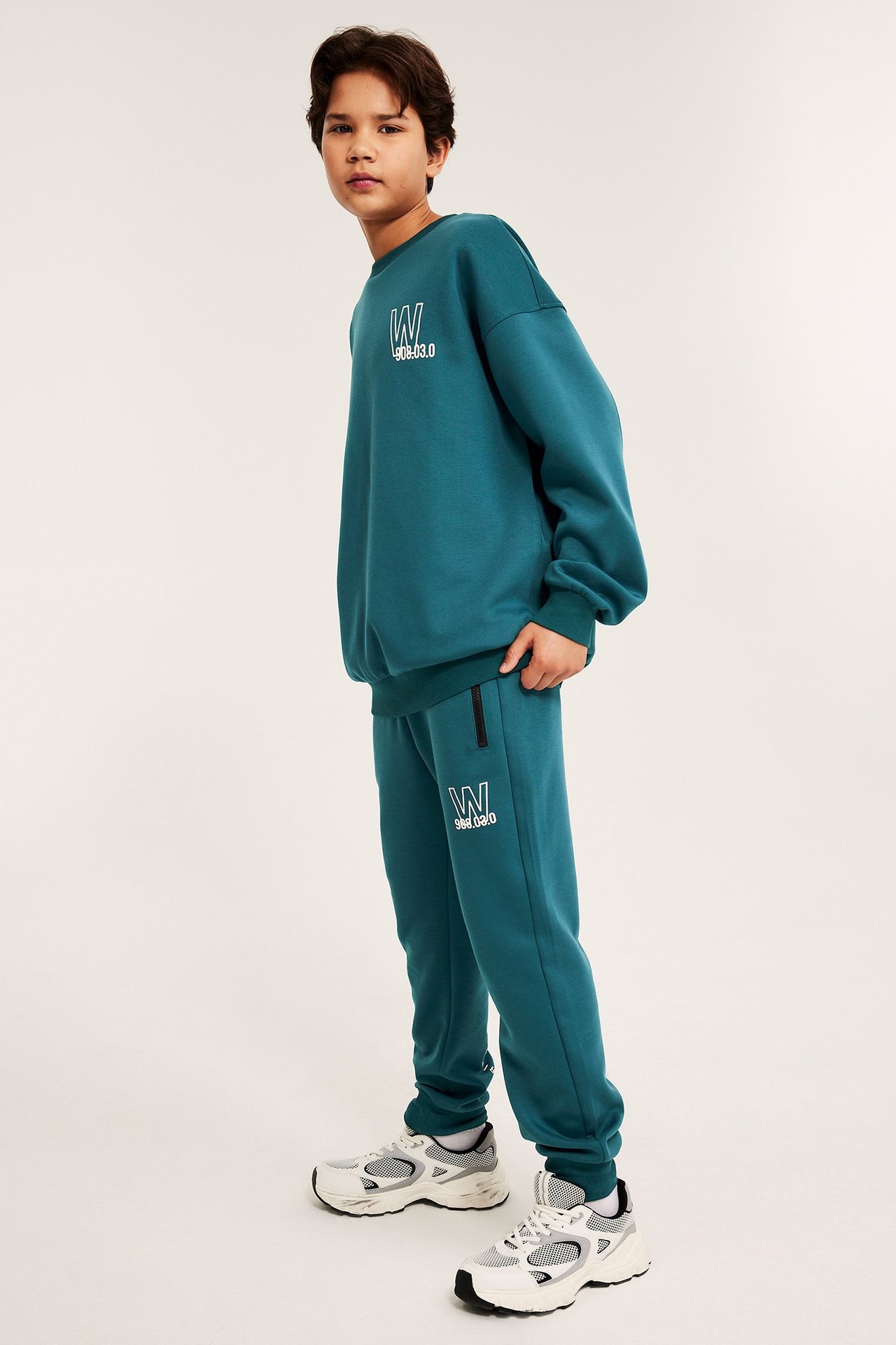 Sivukuva: lapsi vihreissä scuba-collegehousuissa valkoisella painatuksella, vetoketjulla, yhteensopivalla paidalla ja valkoisissa lenkkareissa.