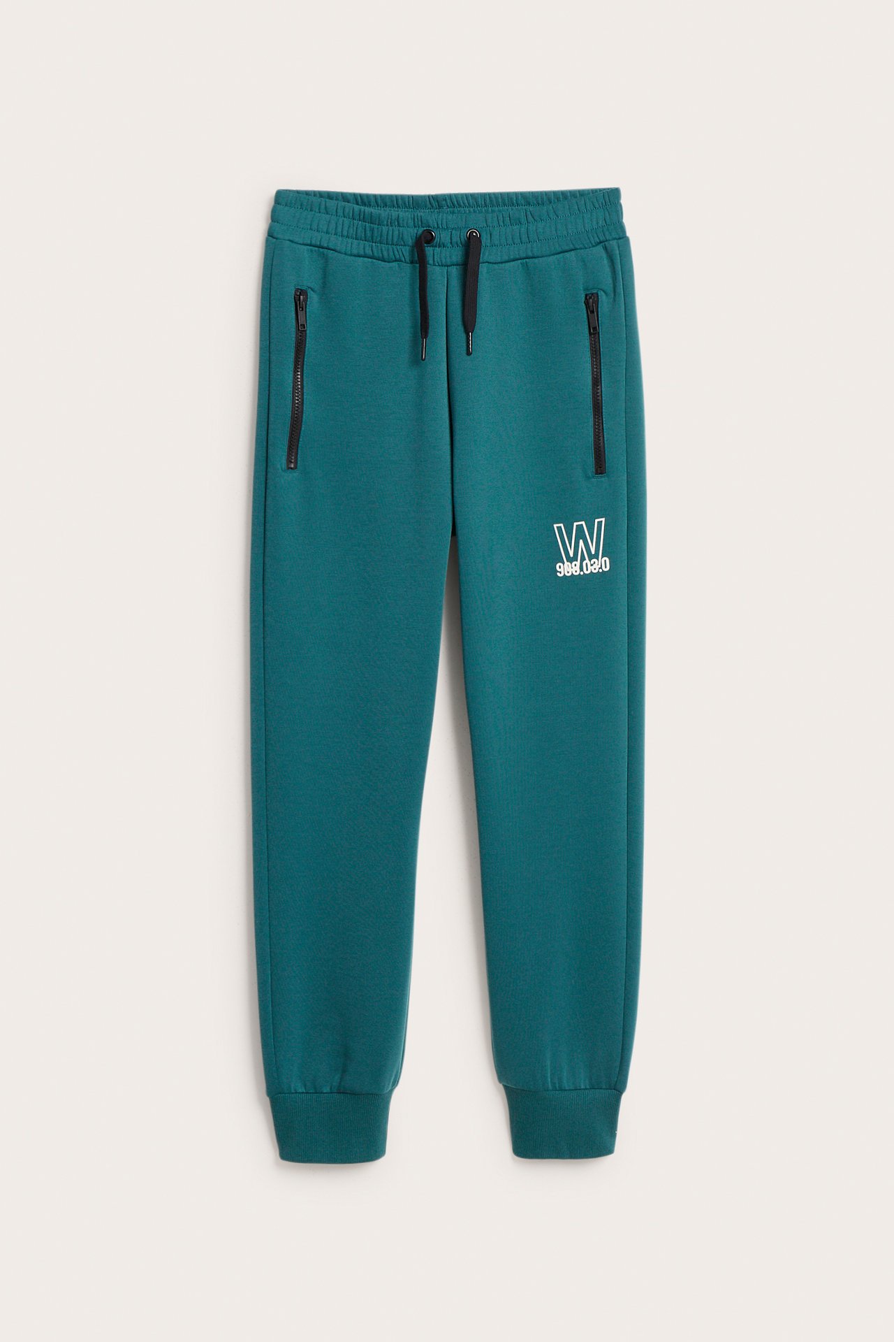 Widok z przodu zielonych spodni dresowych scuba z białym nadrukiem i czarnymi kieszeniami na zamek.