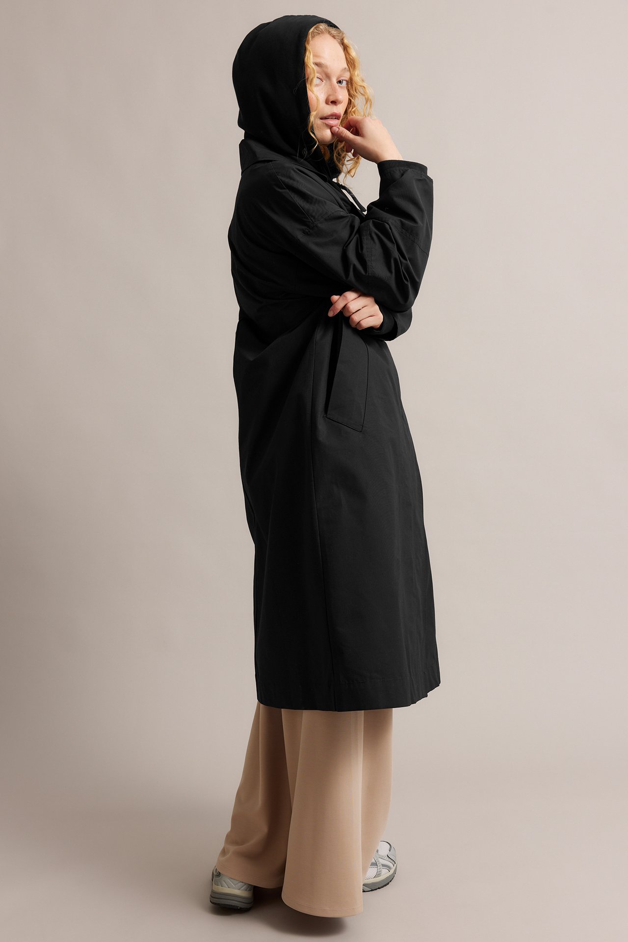 Sidovy: Kvinna i svart huvförsedd trenchcoat av bomullsblandning, beiga vida byxor och grå sneakers.