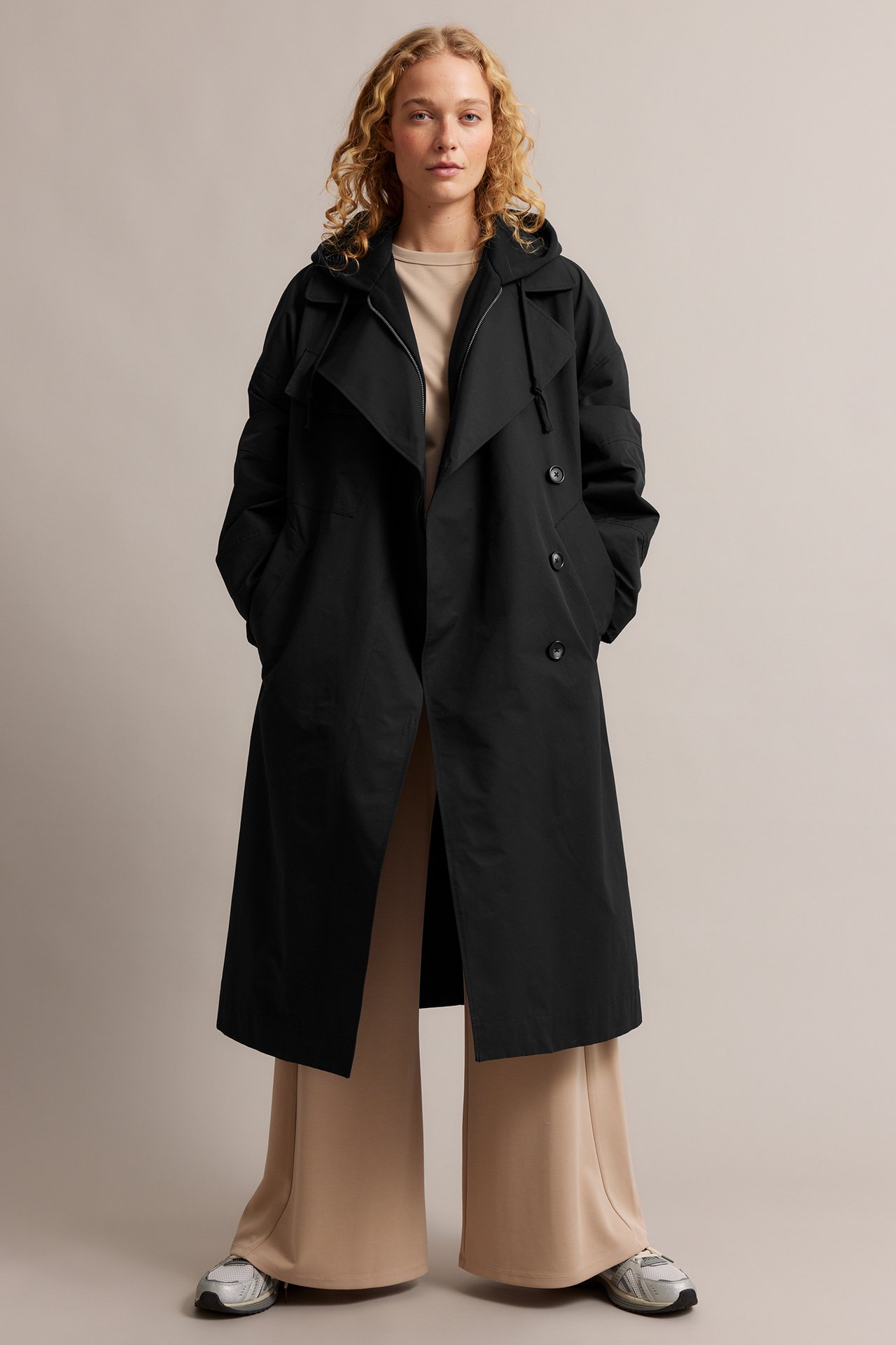 Frontvisning: Kvinne i svart hette-trenchcoat, over beige topp, vide bukser og joggesko.