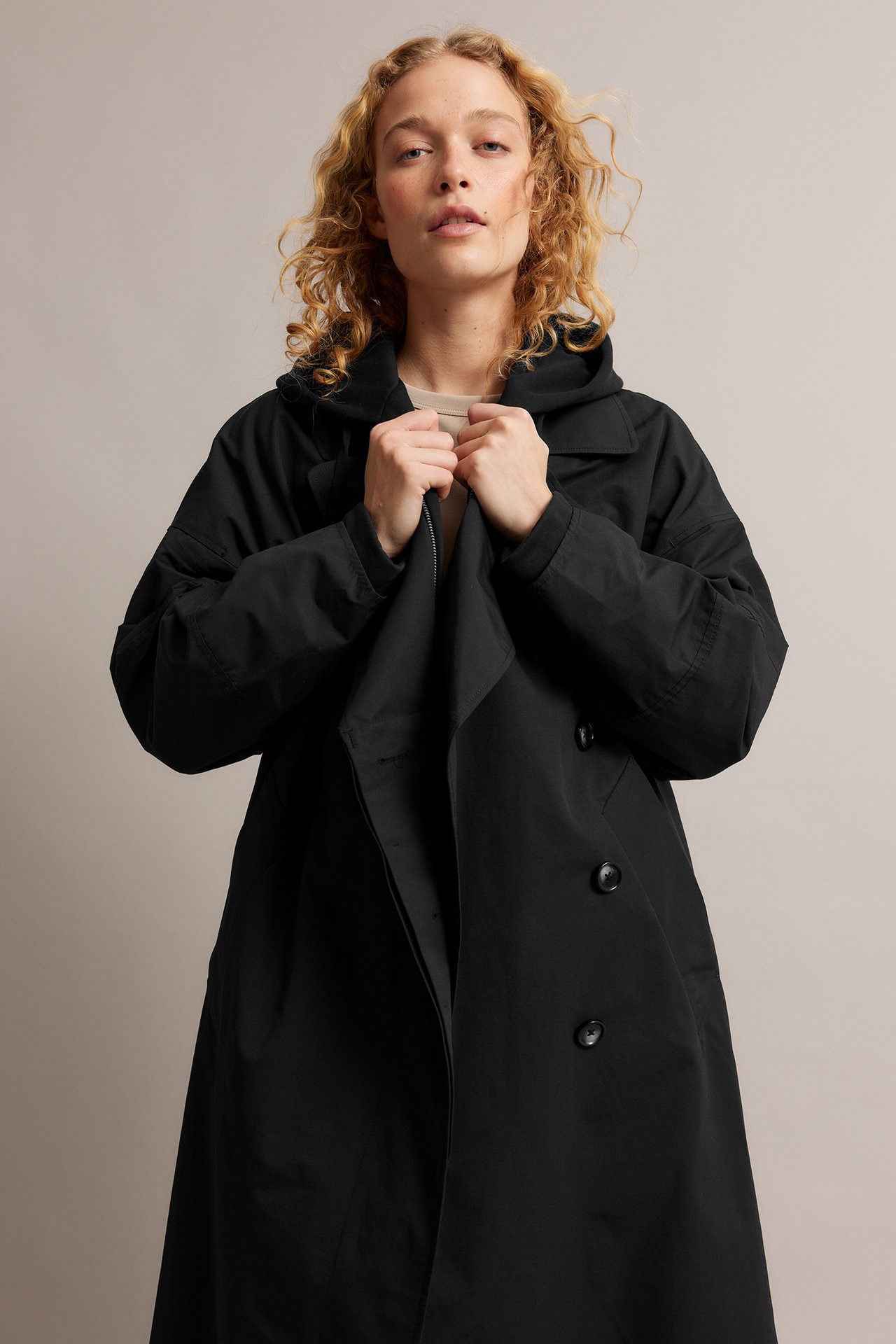 Framifrån: Kvinna i lös svart bomullsblandning trenchcoat med huva, bärs över beige topp.
