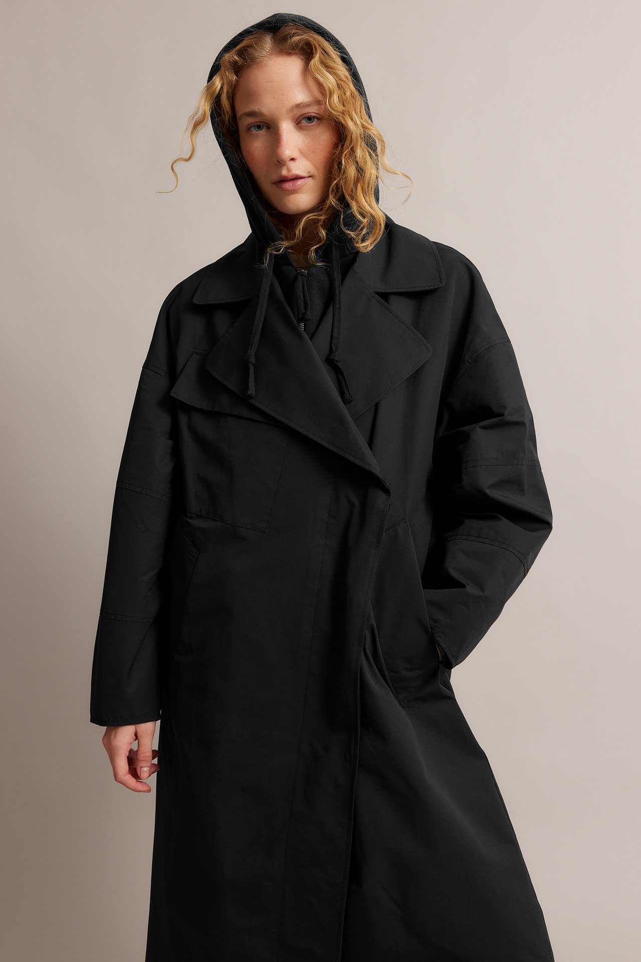 Framifrån, kvinna i svart trenchcoat i bomullsblandning med svart huvtröja under.