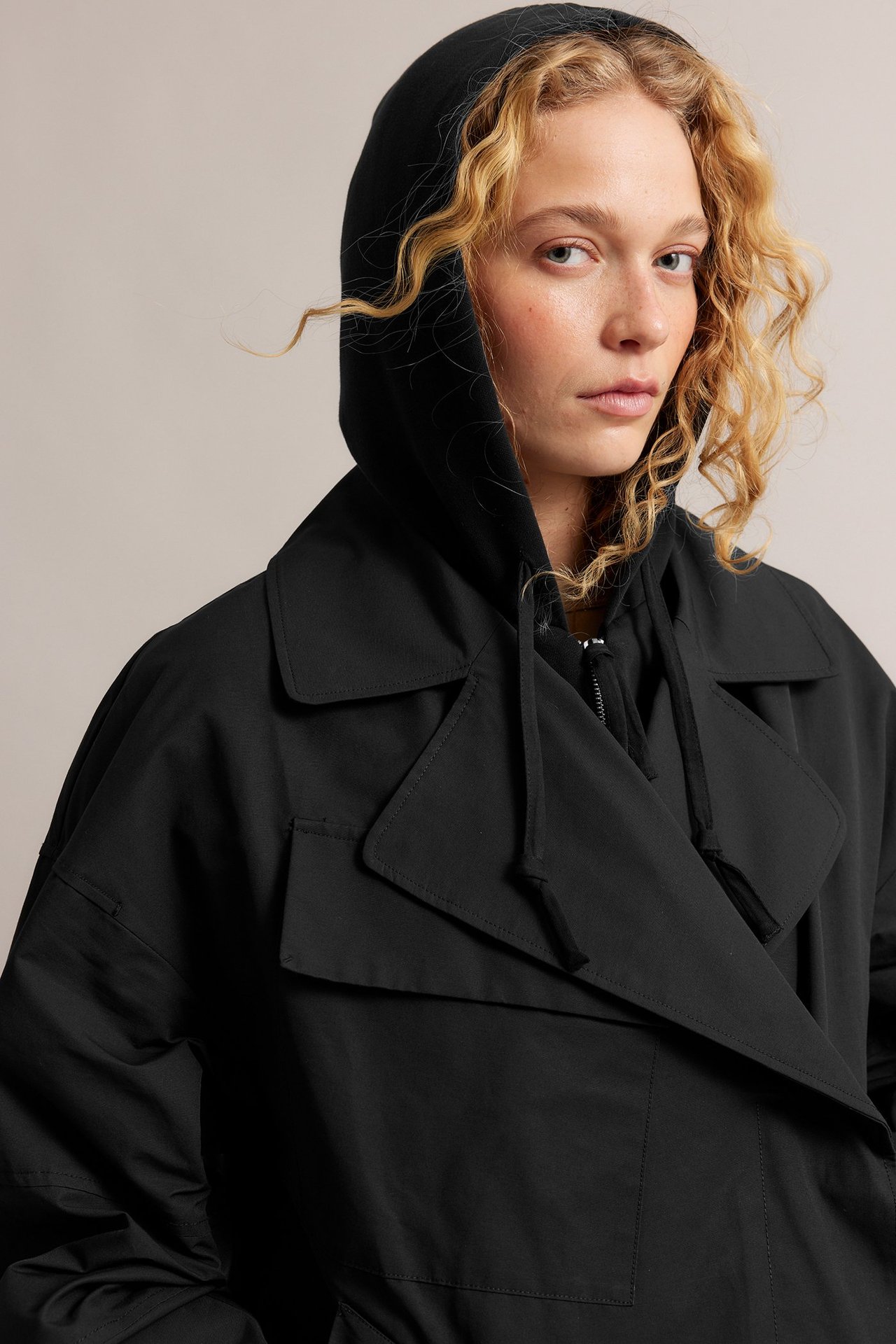 Närbild framifrån på en kvinna i en öppen svart trenchcoat i bomullsblandning över en svart huvtröja.