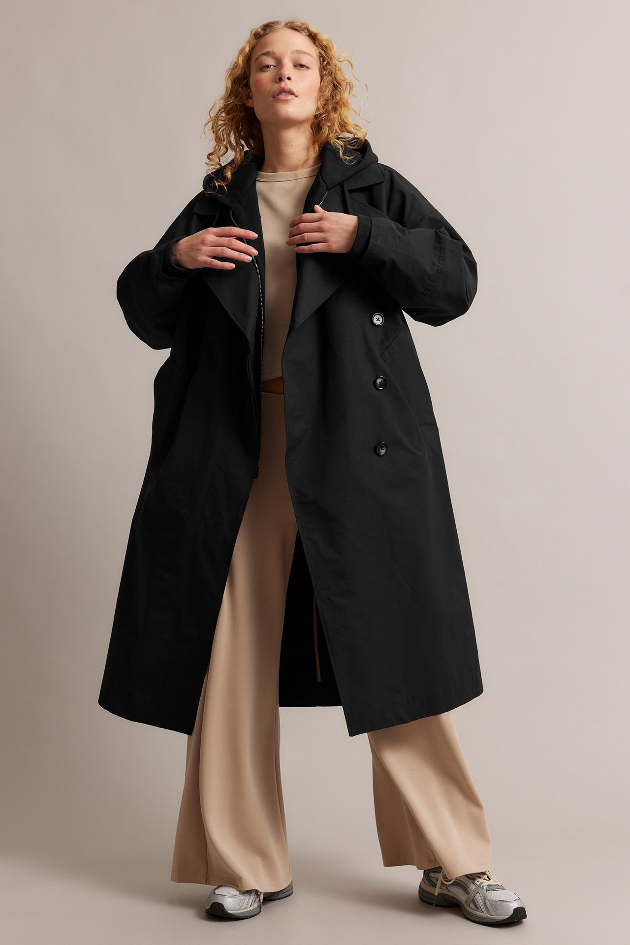 Widok z przodu kobiety w otwartym czarnym trenczu, beżowym topie i szerokich spodniach.