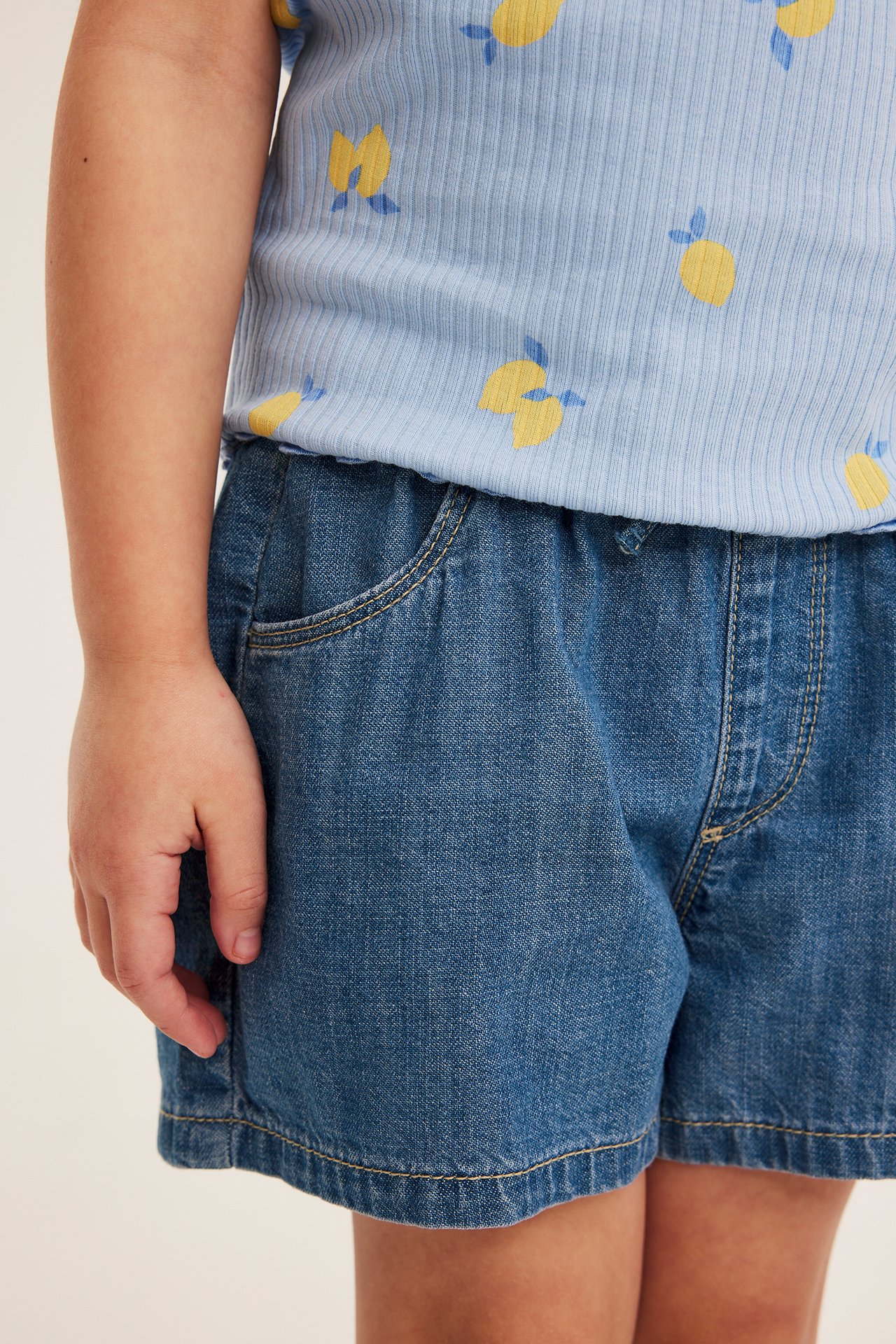 Front view av barns ljusblå dra-på-jeansshorts med fickor, stylade med en citrontryckt topp.