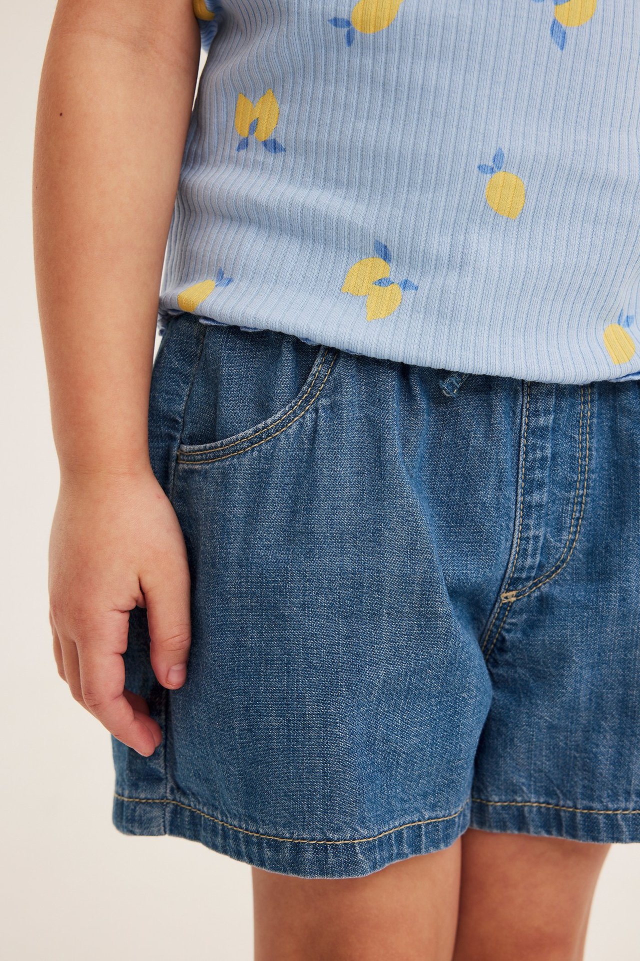 Widok z przodu: dziecięce jasnoniebieskie jeansowe szorty wsuwane z kieszeniami, w zestawie z bluzką w cytryny.
