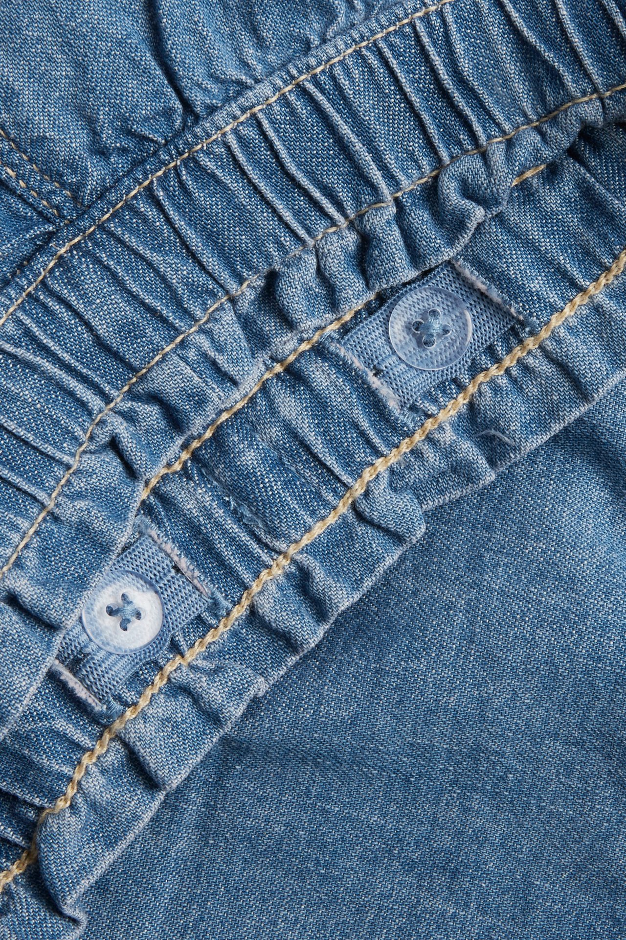 Zbliżenie na jasnoniebieskie szorty jeansowe, z elastyczną, marszczoną talią, dwoma jasnoniebieskimi guzikami i beżowymi przeszyciami.