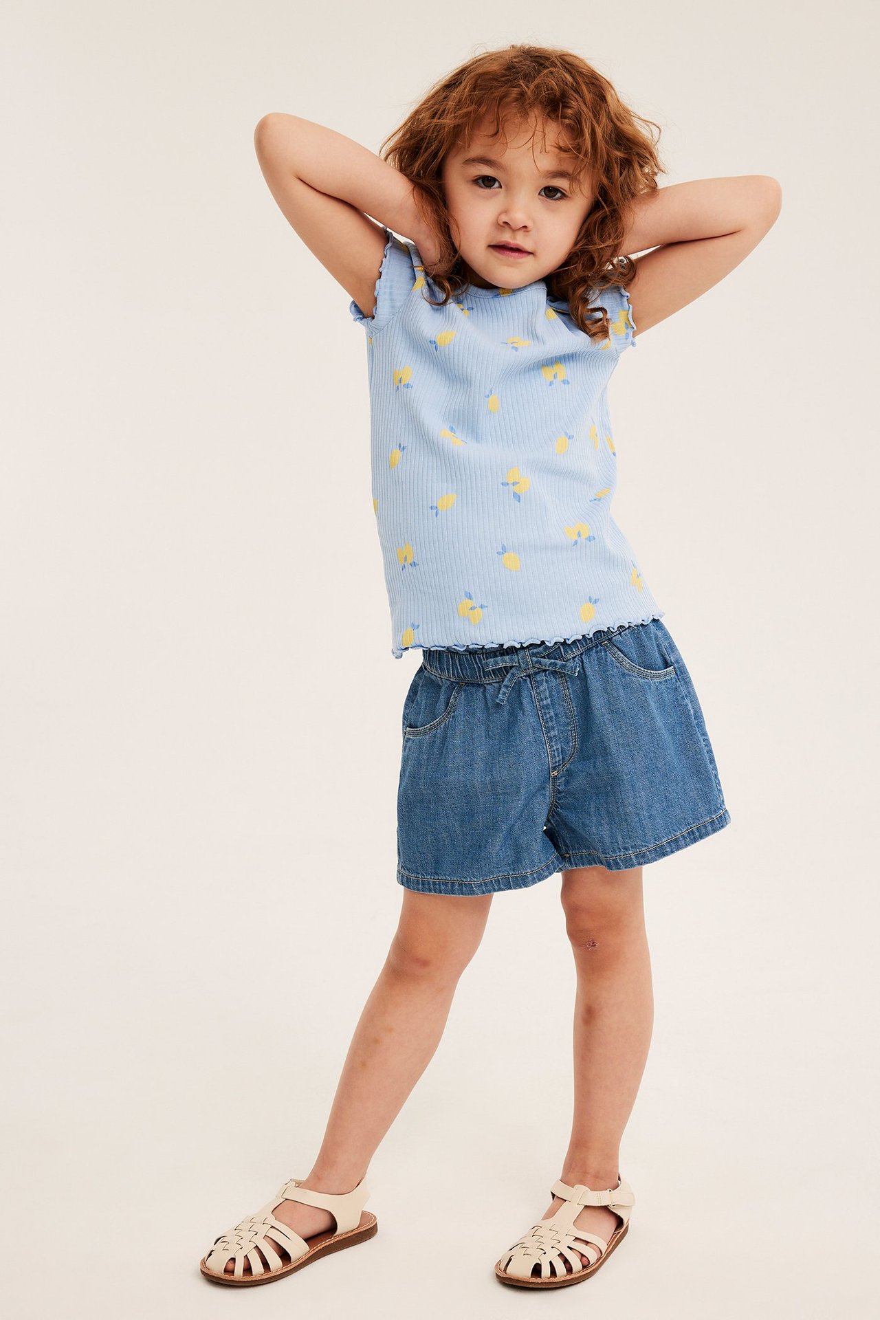 Framifrån, ett barn i ljusa denim dra-på-shorts, en ljusblå ribbad t-shirt med citroner och beiga sandaler.