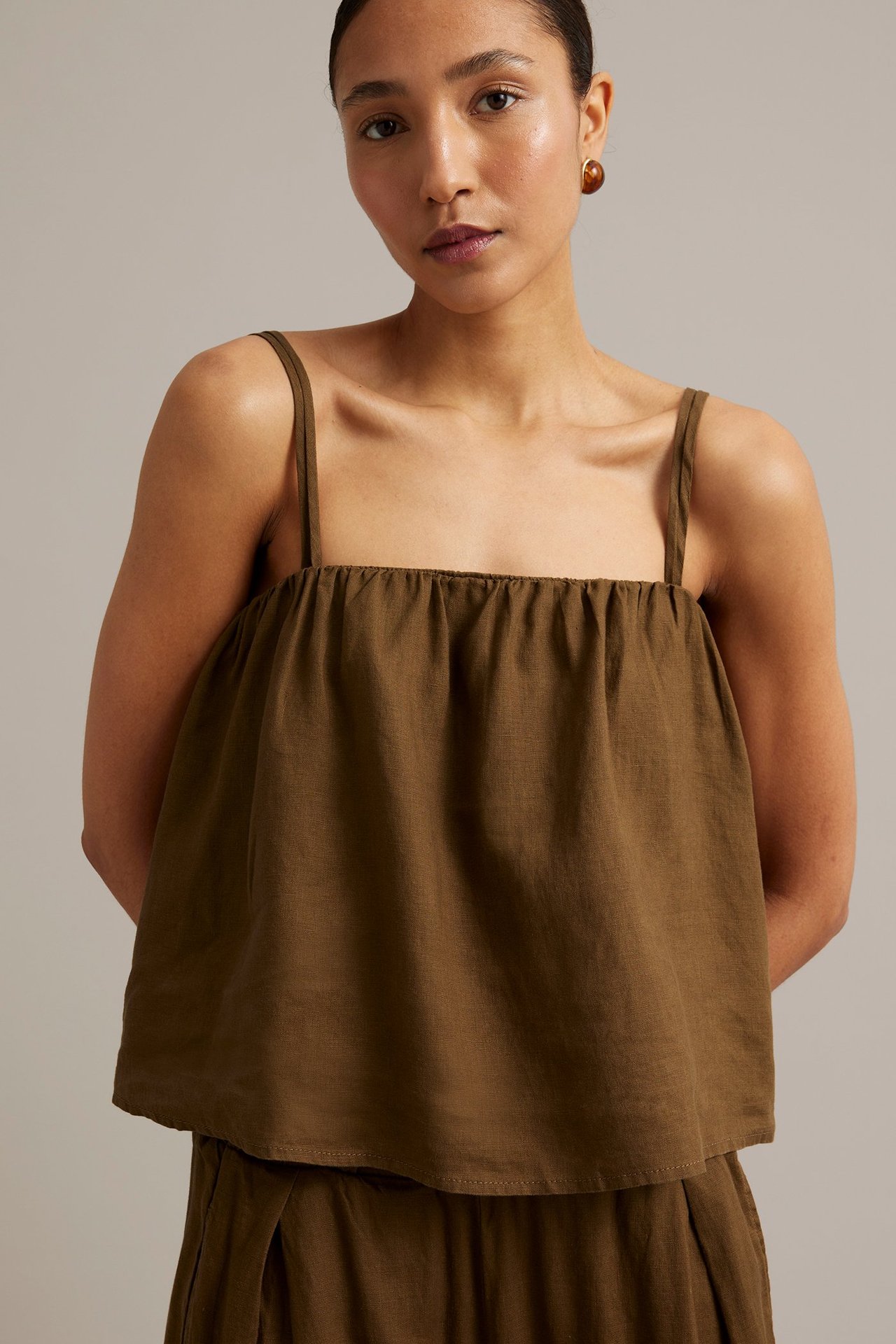 Framifrån, kvinna i brunt linne med smala axelband, bruna örhängen och matchande byxor.