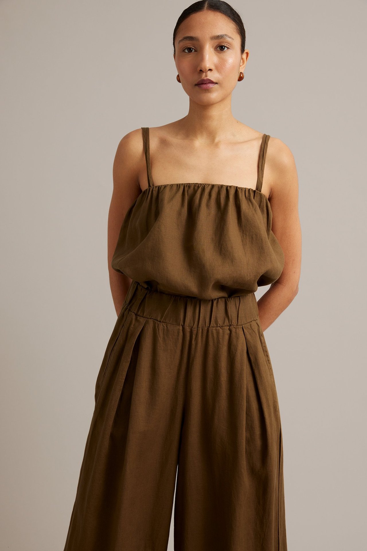 Framifrån, kvinna i brunt linne med smala axelband, matchande vida linnebyxor och små bruna örhängen.
