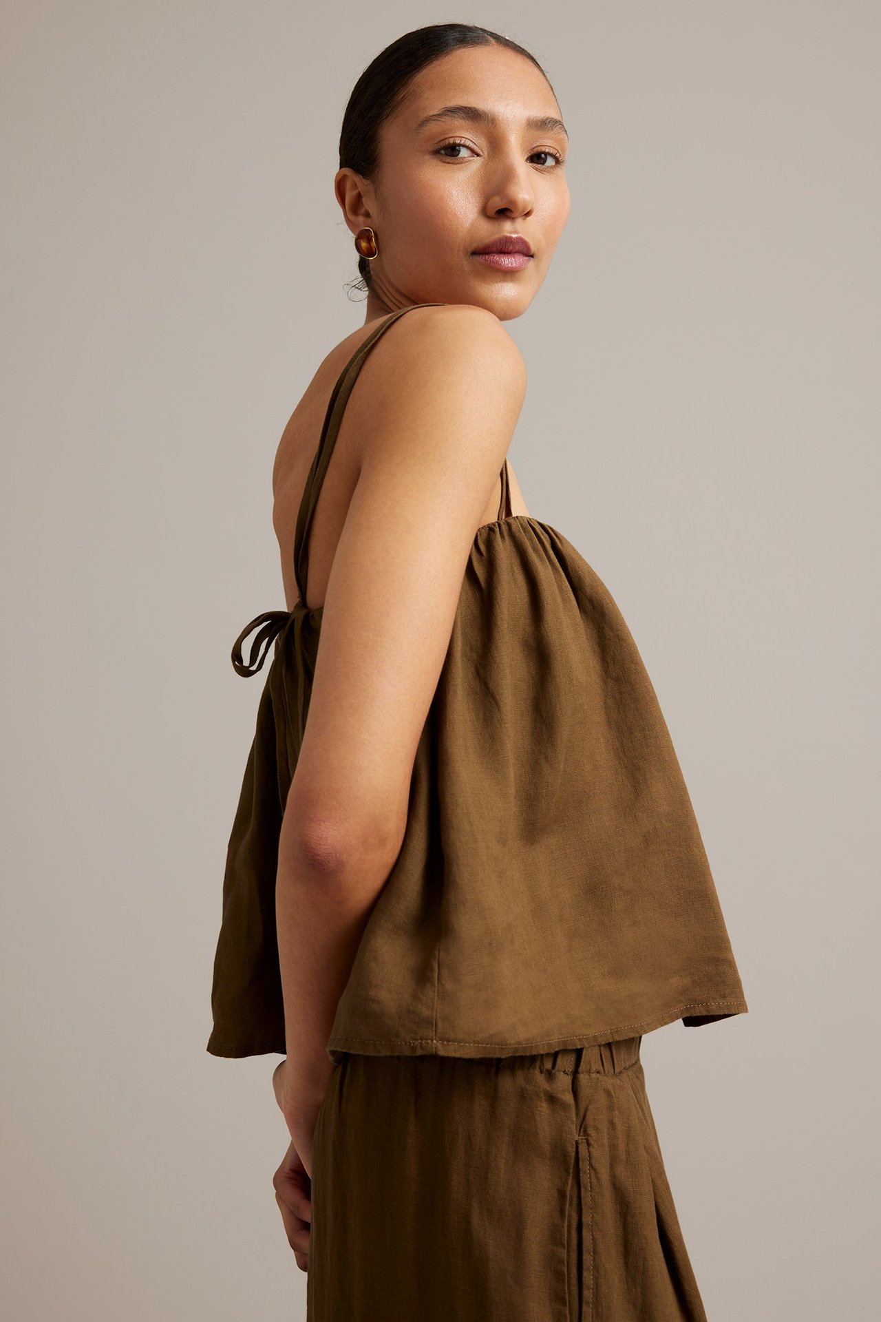 Kvinna i brunt linne av linne med smala axelband, knytband bak och bruna ovala örhängen.
