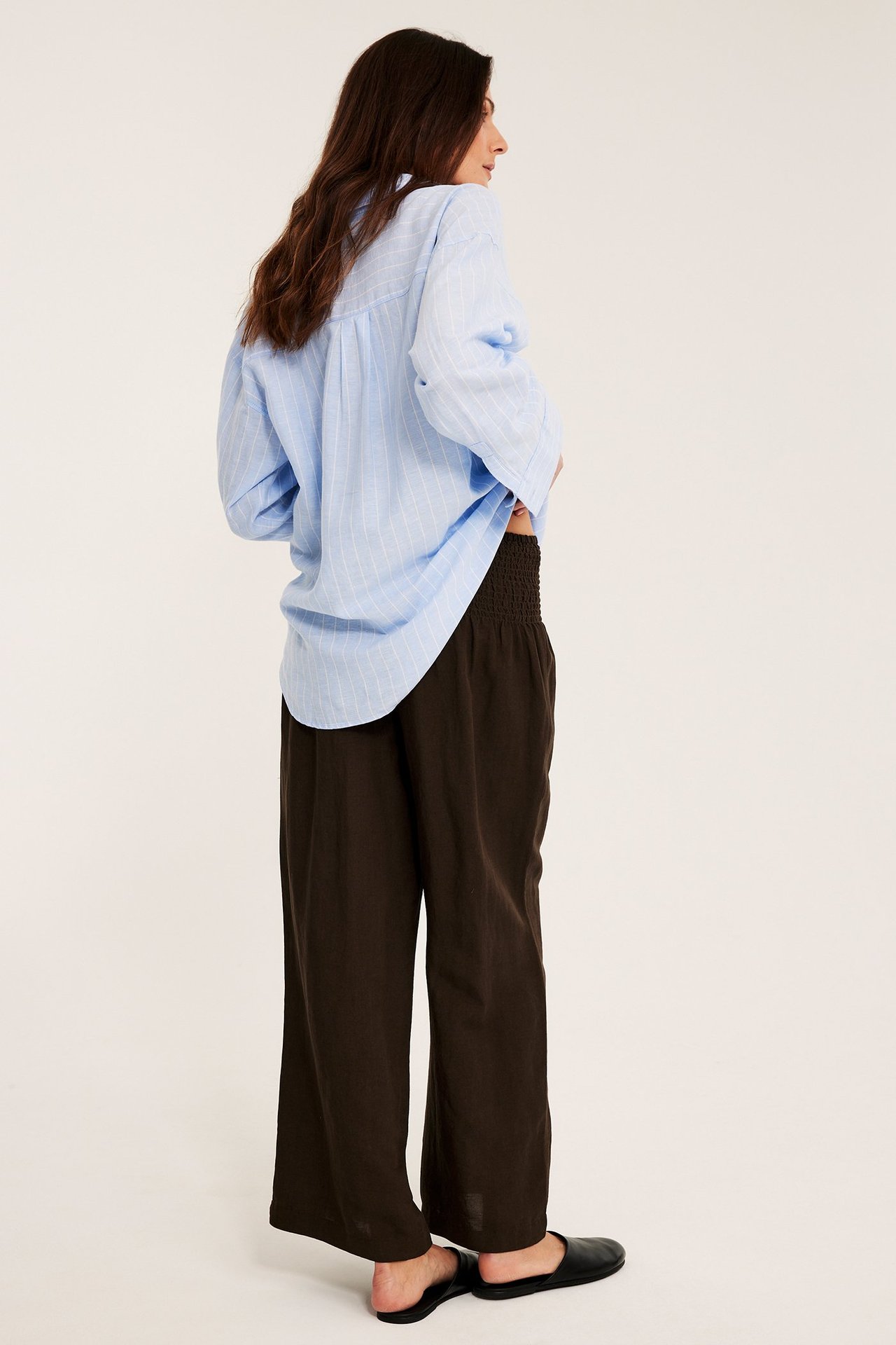 Sidebilde av en kvinne i mørkebrune vide linbuks med smock i midjen og en lyseblå stripete skjorte.