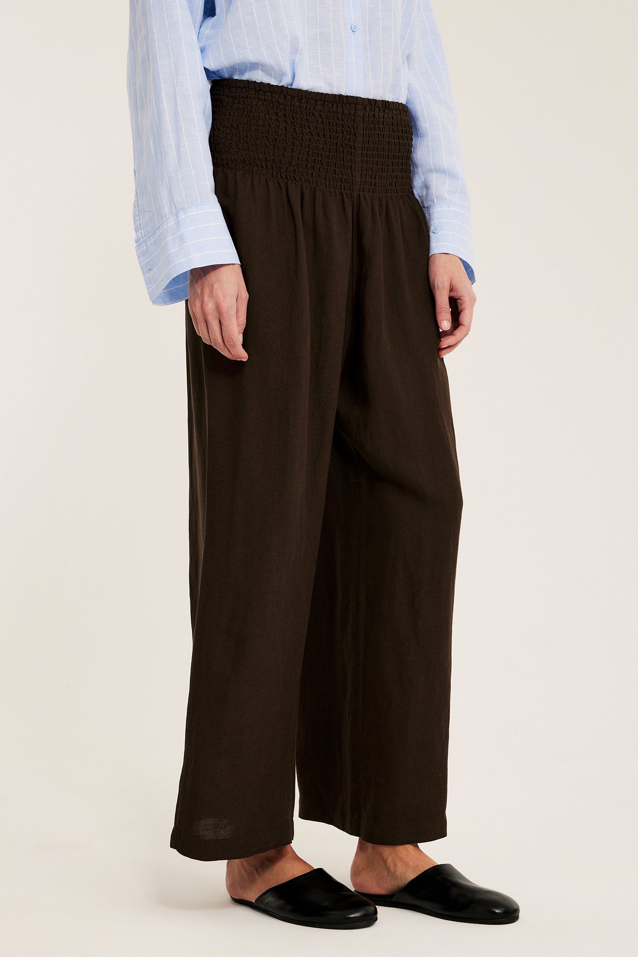 Sidebilde: Kvinne i mørkebrune vide linblandingsbukser med smock, lyseblå stripet skjorte, svarte sko.