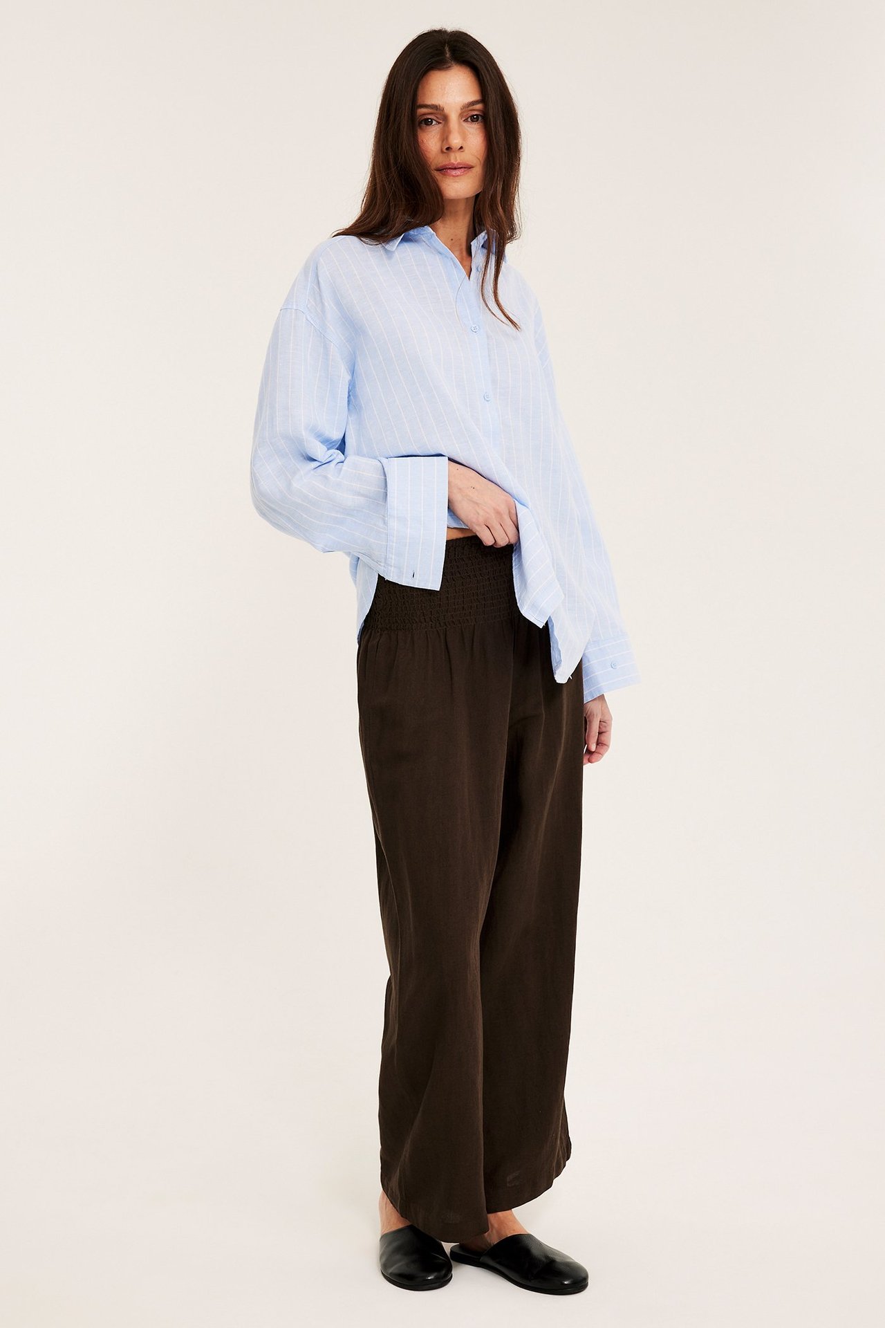 Kvinne i mørkebrune vide linbuksermed smocksøm i midjen, lyseblå stripete skjorte og svarte mules.