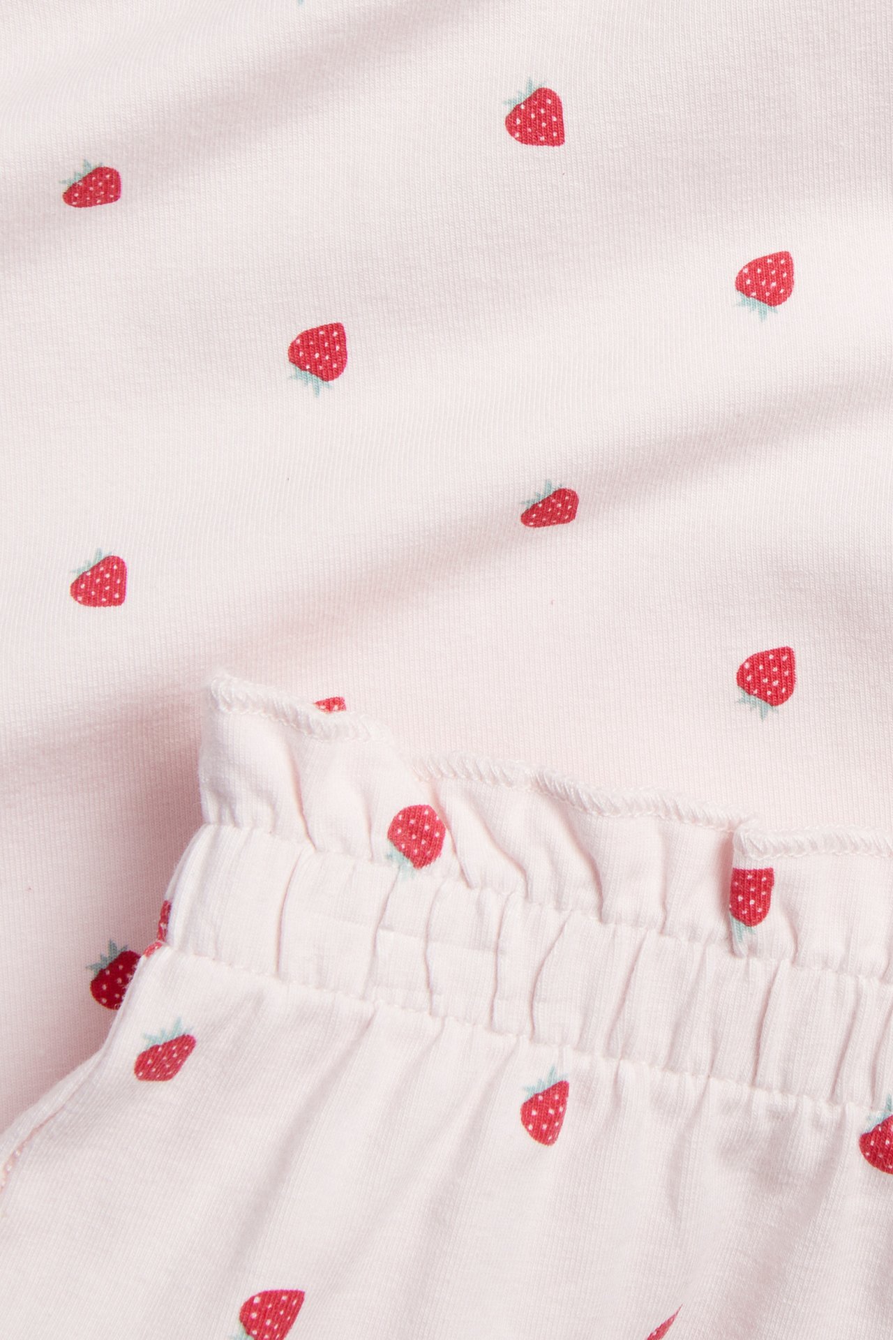 Nærbilde av lysrosa bomullspyjamas med rødt jordbærmønster og volangkantet, rynket linning.