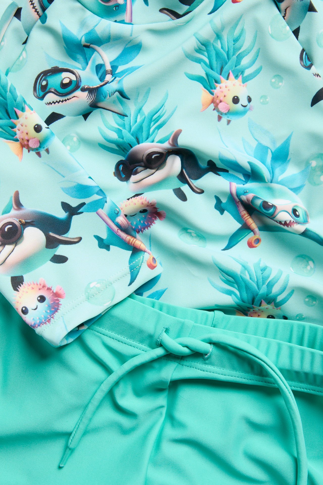 Nærbilde av et lysturkis UV-sett for barn; mønstret topp med haier og ensfargede shorts med snøring.