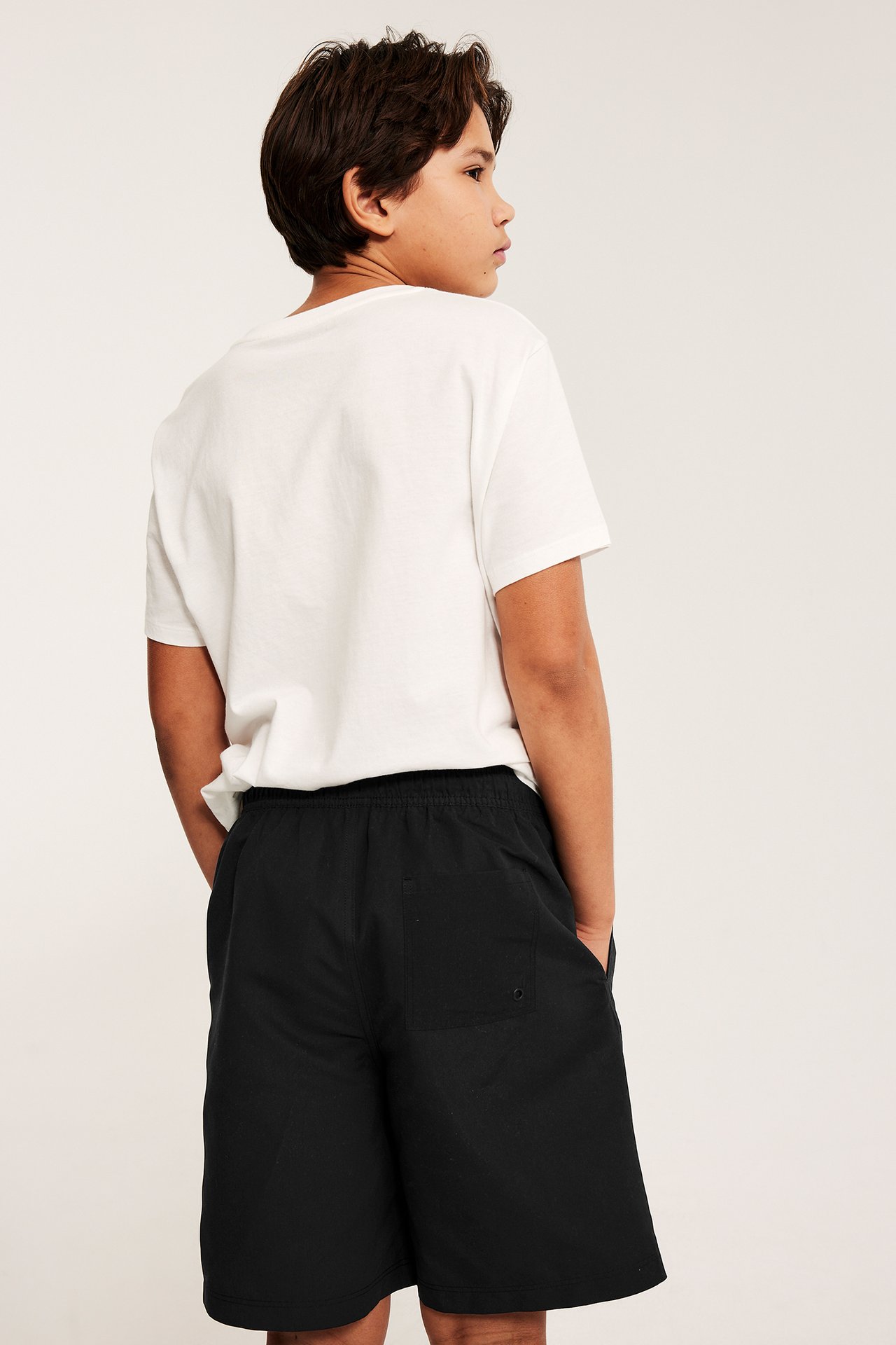 Takakuva: Lapsi käyttää yksivärisiä mustia uimashortseja takataskulla ja valkoista t-paitaa.