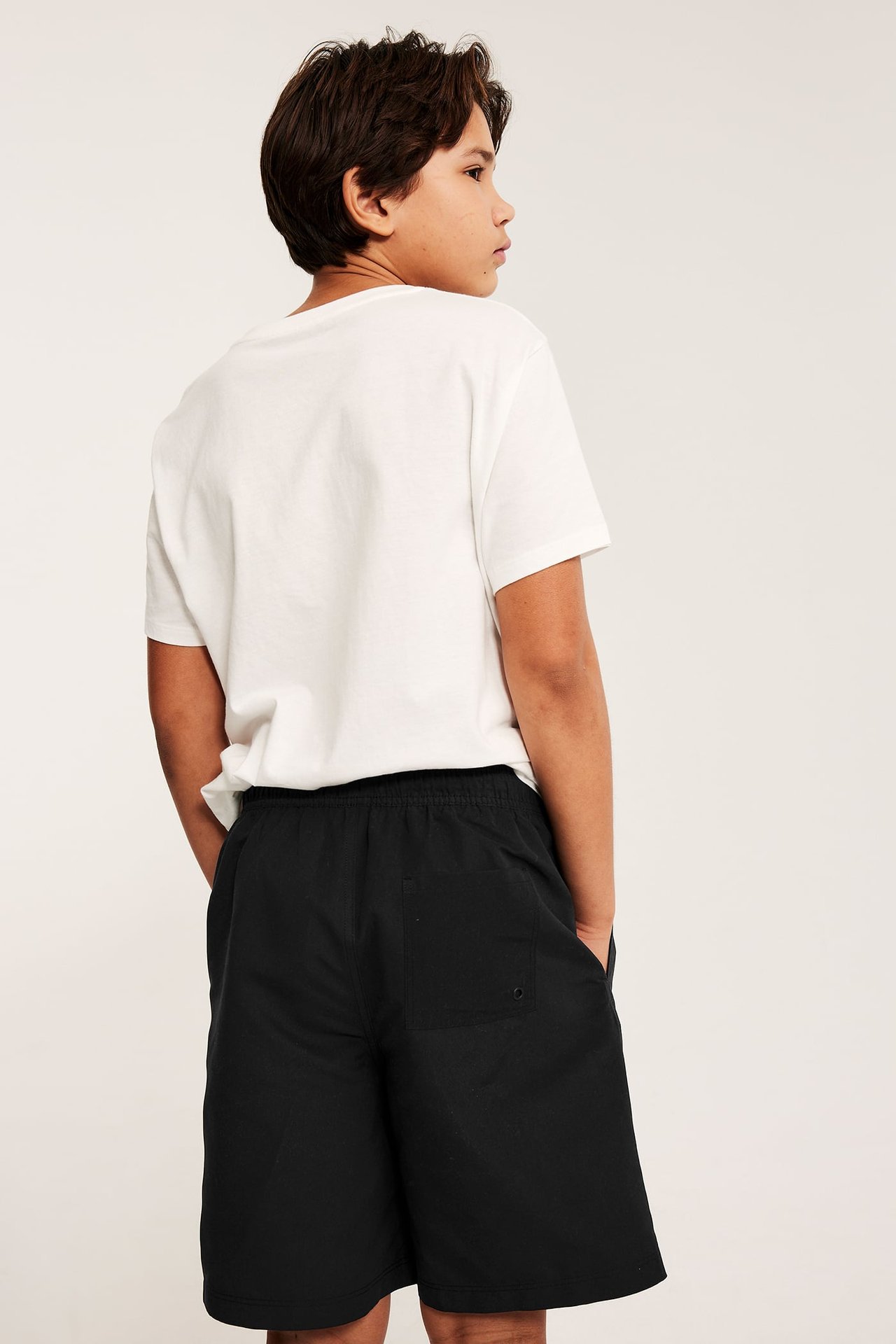 Bakifrån: Ett barn bär enfärgade svarta badshorts med bakficka och en vit t-shirt.