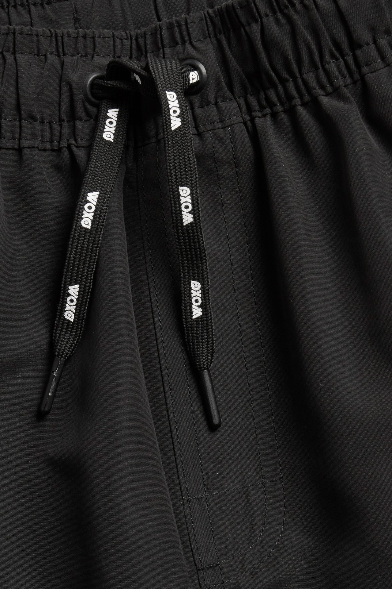 Lähikuva mustista uimashortseista, joissa resori ja musta, valkoisella "WOXG"-tekstillä varustettu kiristysnyöri.