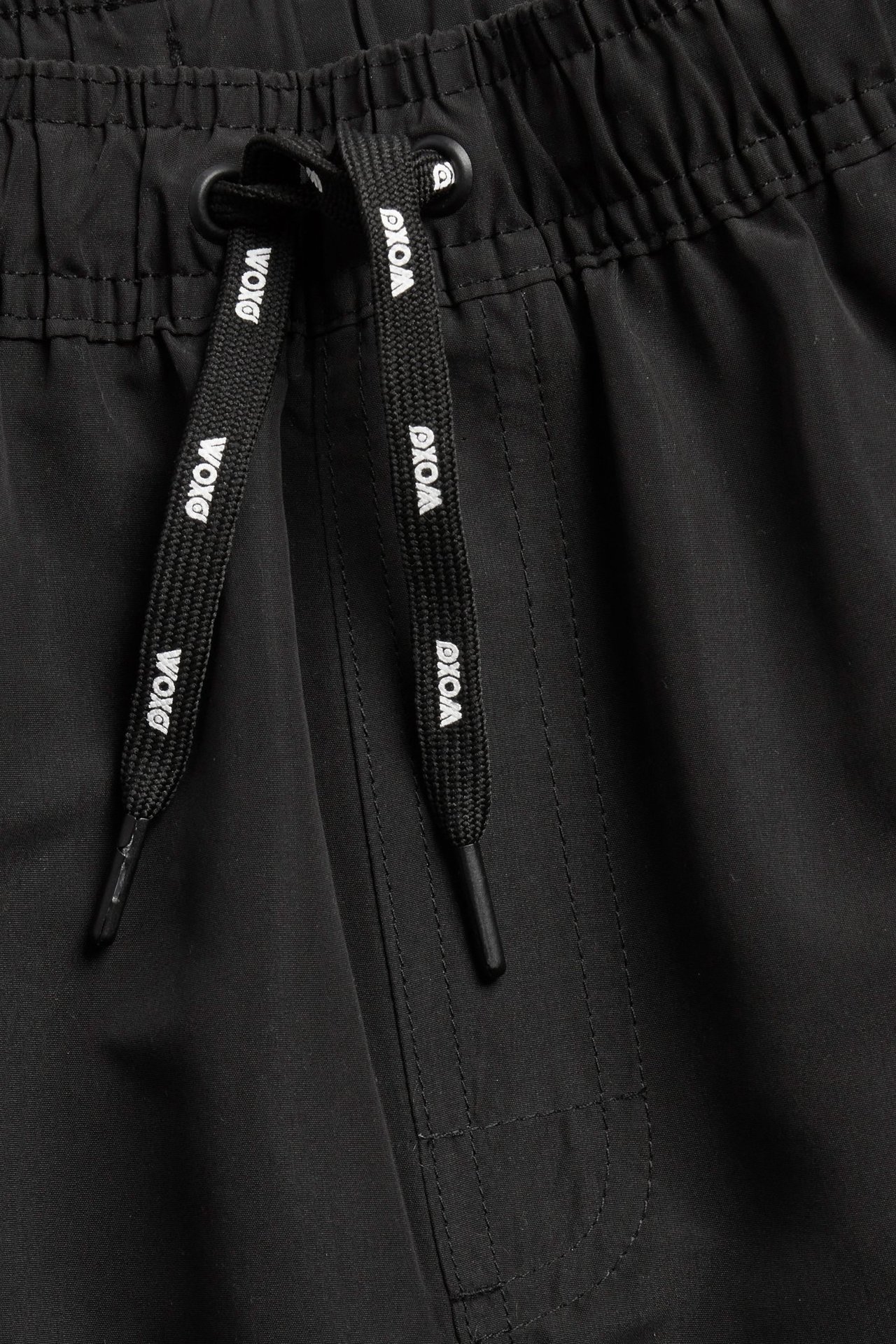 Lähikuva mustista uimashortseista, joissa resori ja musta, valkoisella "WOXG"-tekstillä varustettu kiristysnyöri.