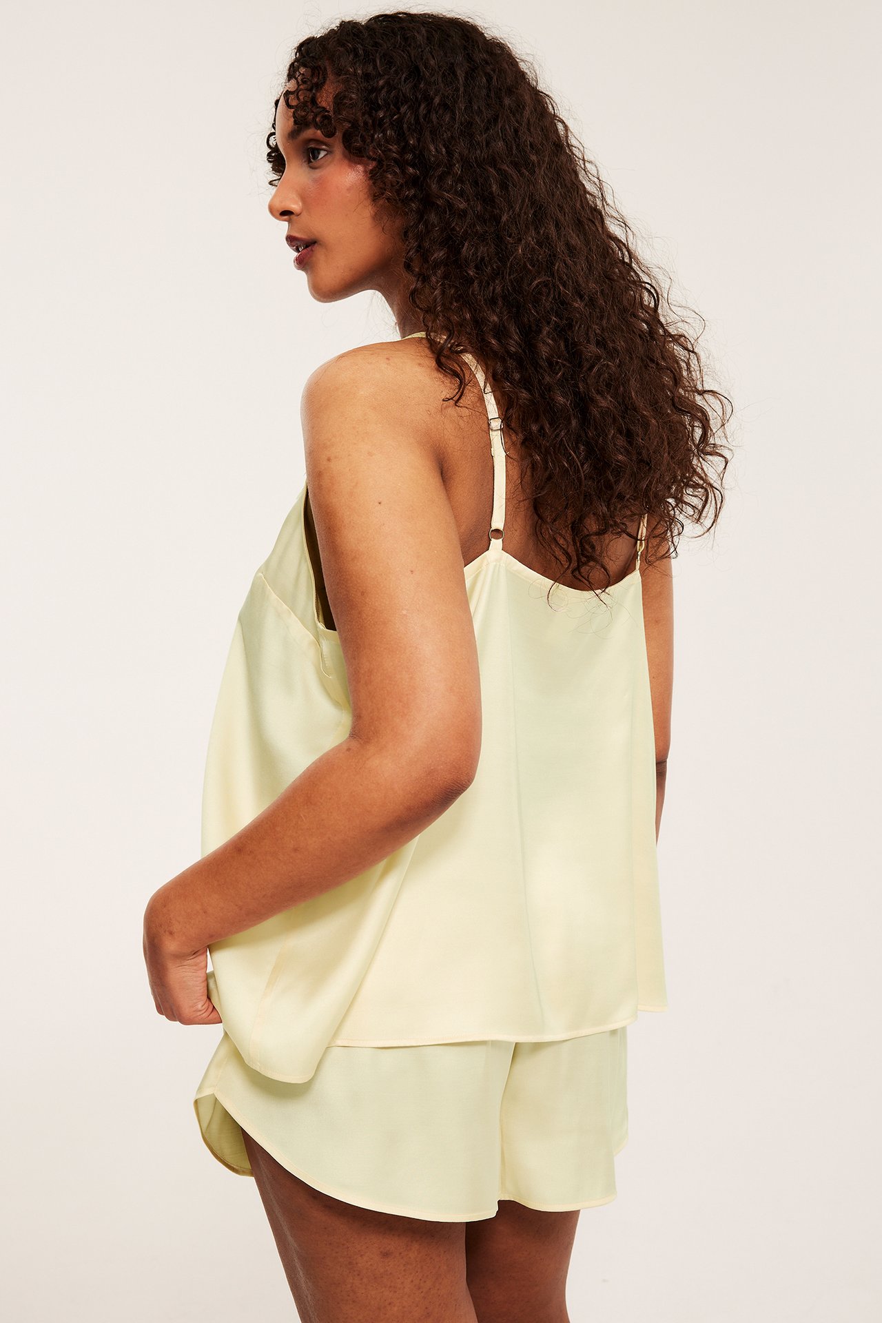 Sidovy av en kvinna i ljusgul satinpyjamas, en camisole med reglerbara band och matchande shorts.