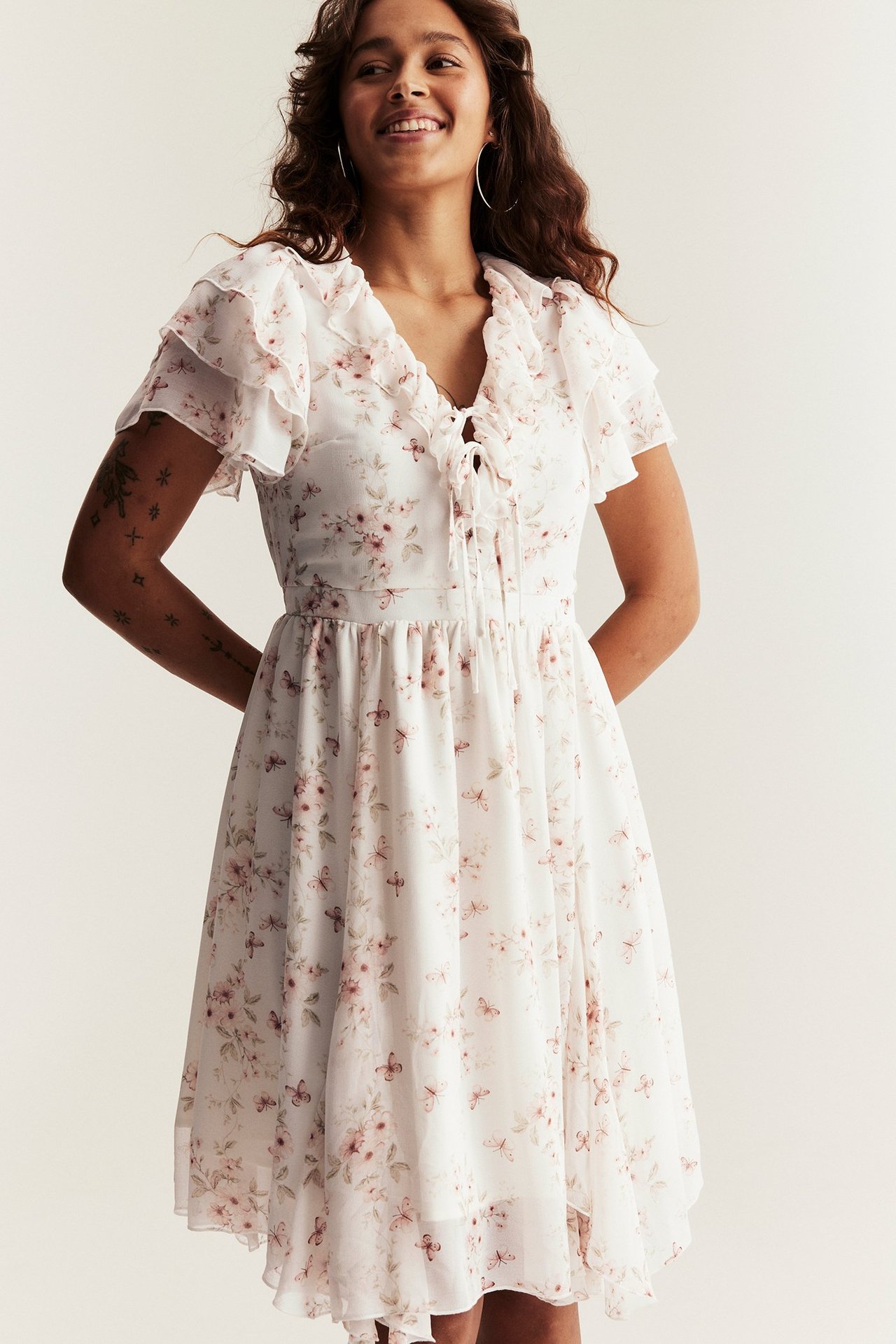 Framsida: kvinna i off-white blommig chiffongklänning med volanger, knytband fram, korta ärmar, ringörhängen.