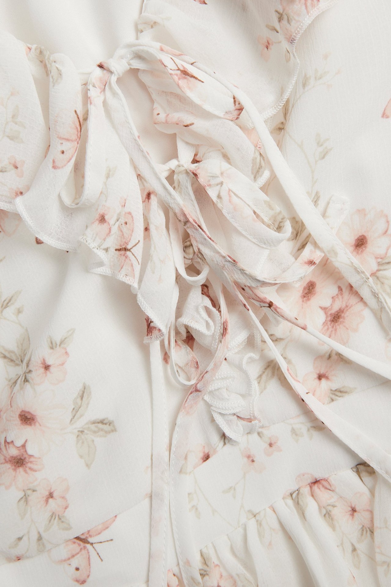Nærbilde av en offwhite chiffonkjole med rosa blomstermønster, knytebånd foran og volanger.
