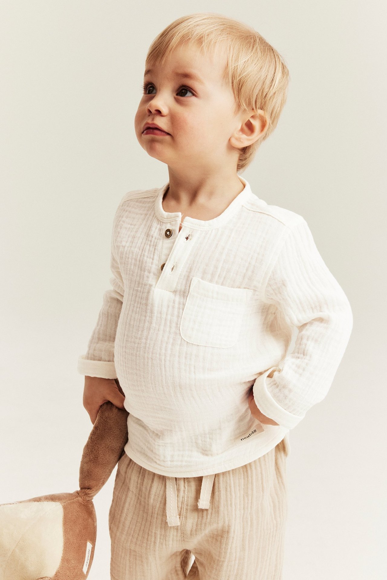 Barn i offwhite långärmad muslin topp med knappar, ficka; beiga byxor; håller ett gosedjur.