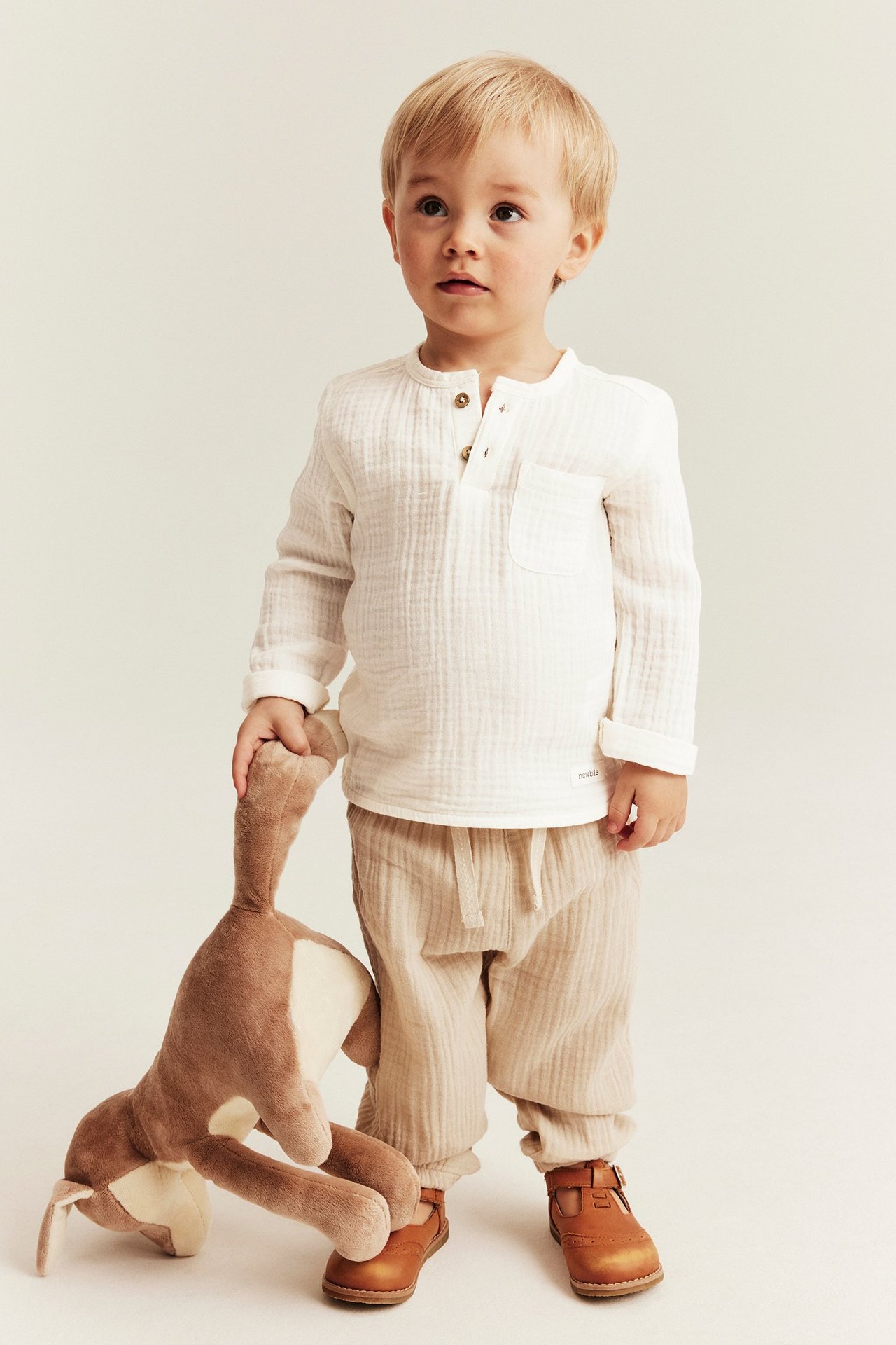 Dziecko nosi kremowy muślinowy top z długim rękawem, beżowe spodnie i brązowe buty, trzymając pluszową zabawkę.