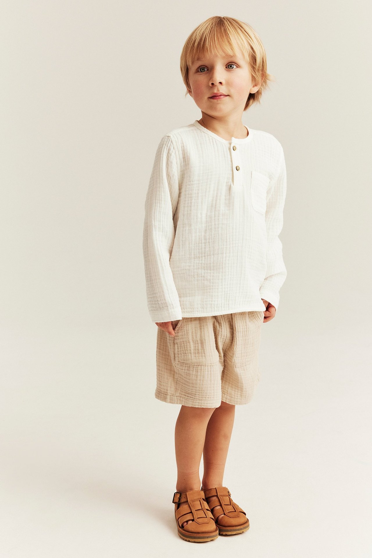 Front view: Kid i benvit långärmad topp i muslin med knappar, beige shorts och bruna sandaler.