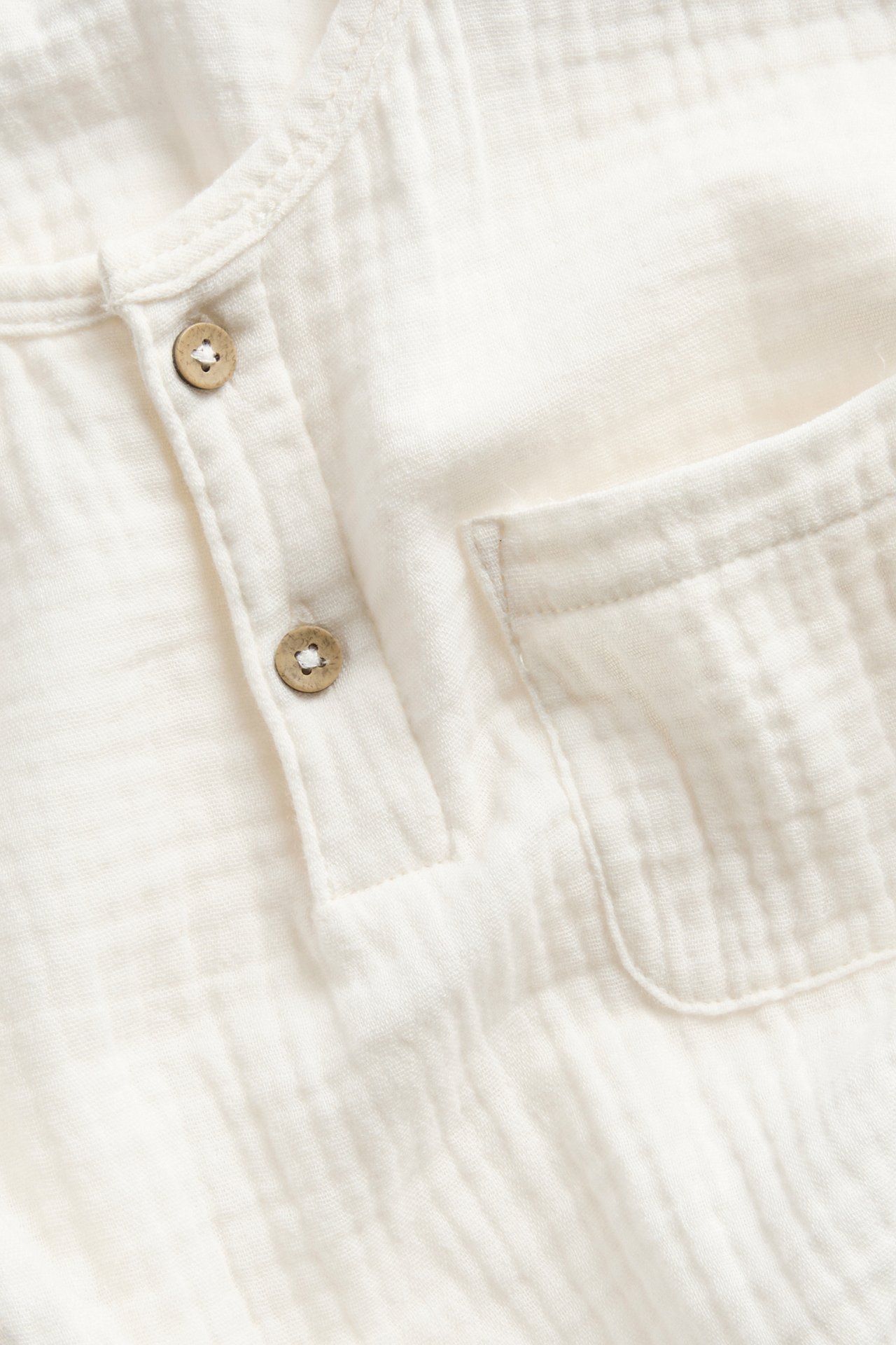 Lähikuva luonnonvalkoisesta puuvillamusliinisesta pitkähihaisesta topista kahdella napilla ja rintataskulla.