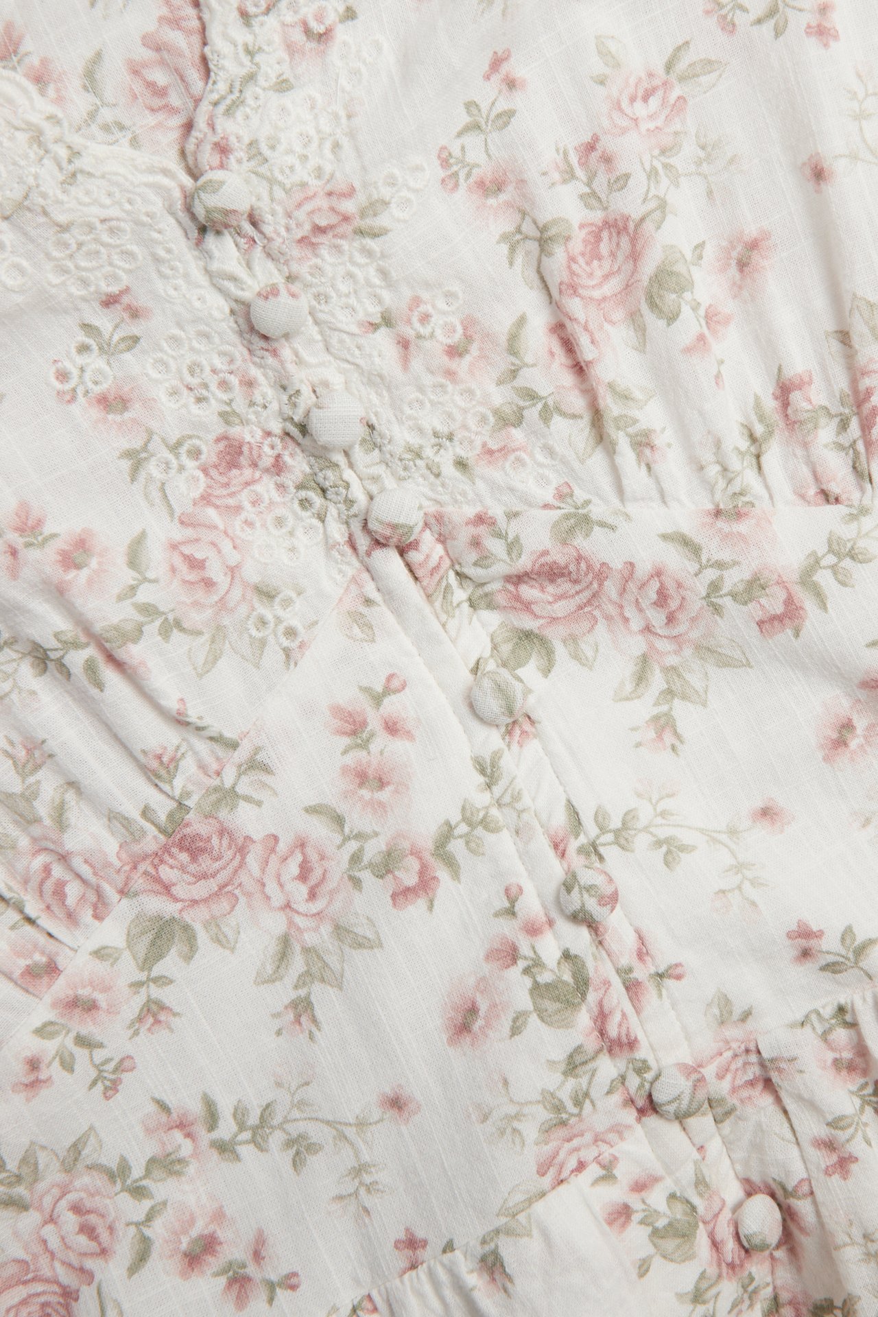 Lähikuva luonnonvalkoisesta puuvillaisesta kukkamekosta, jossa on vaaleanpunaisia ruusuja, kirjailuja ja päällystettyjä nappeja.