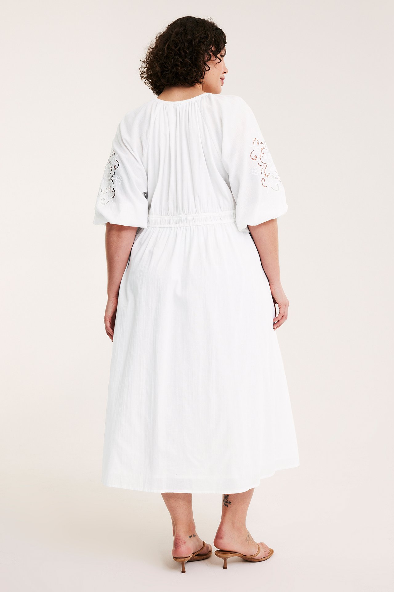 Bakifrån: kvinna i vit bomullsklänning med broderade ärmar, bärande ljusbruna sandaler.