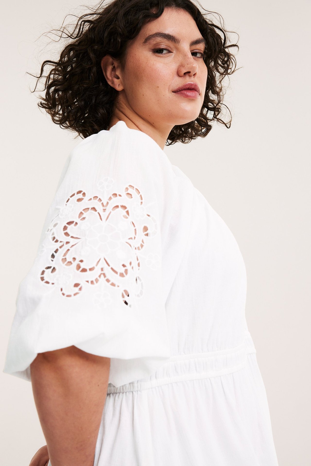 Sidebilde av en kvinne i en hvit bomullskjole med broderte puffermer og elastisk midje.