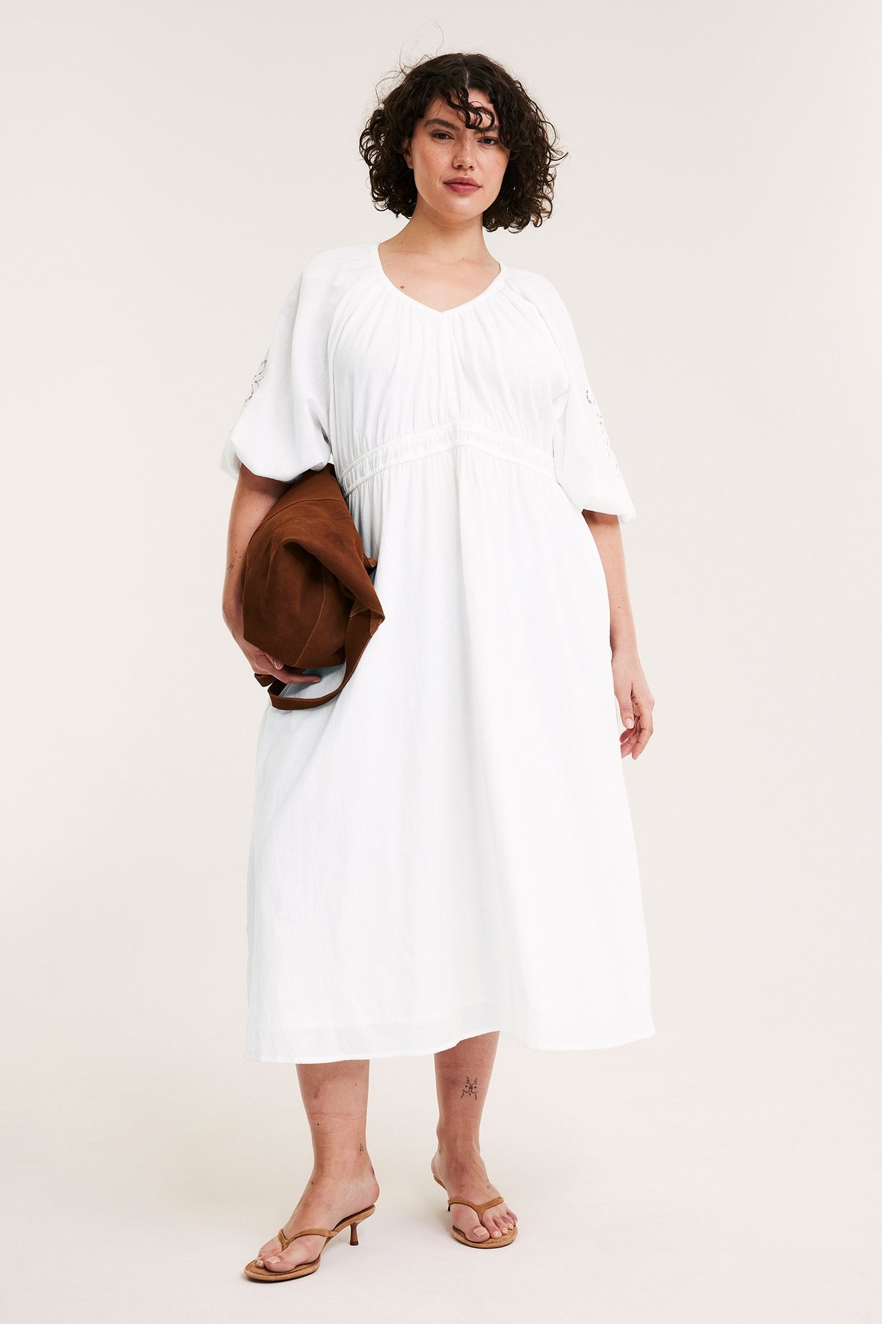 Forfra: kvinne i hvit bomulls V-hals lang kjole med broderte ermer, holder brun veske, brune sandaler.