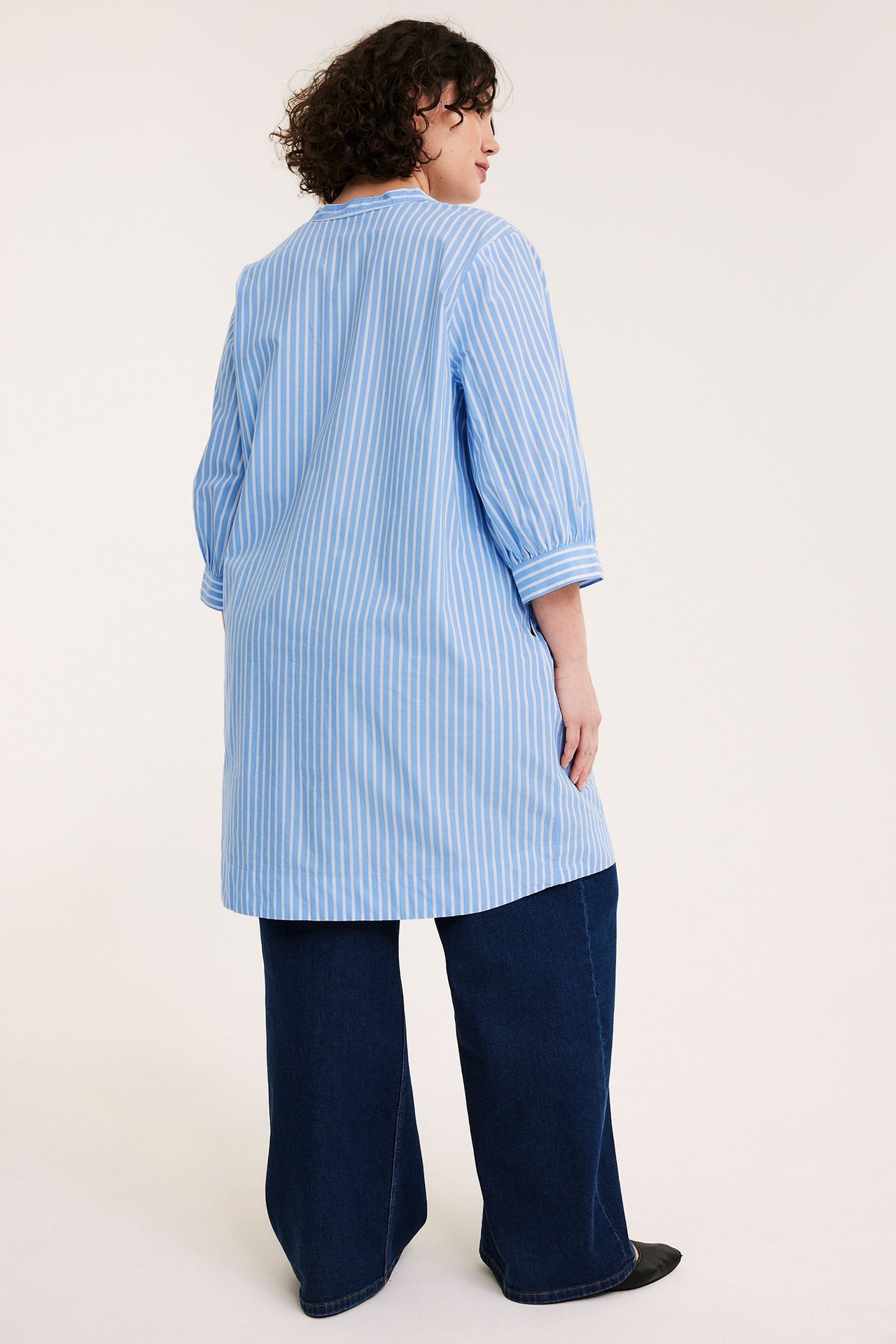 Bakifrån: kvinna i ljusblårandig poplintunika med 3/4-ärm, stylad över mörkblå vida byxor.