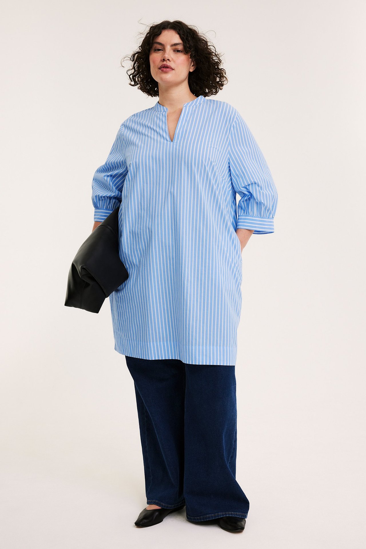 Framifrån: Kvinna i blåvitrandig poplinklänning, mörkblå jeans, svarta skor, håller svart väska.