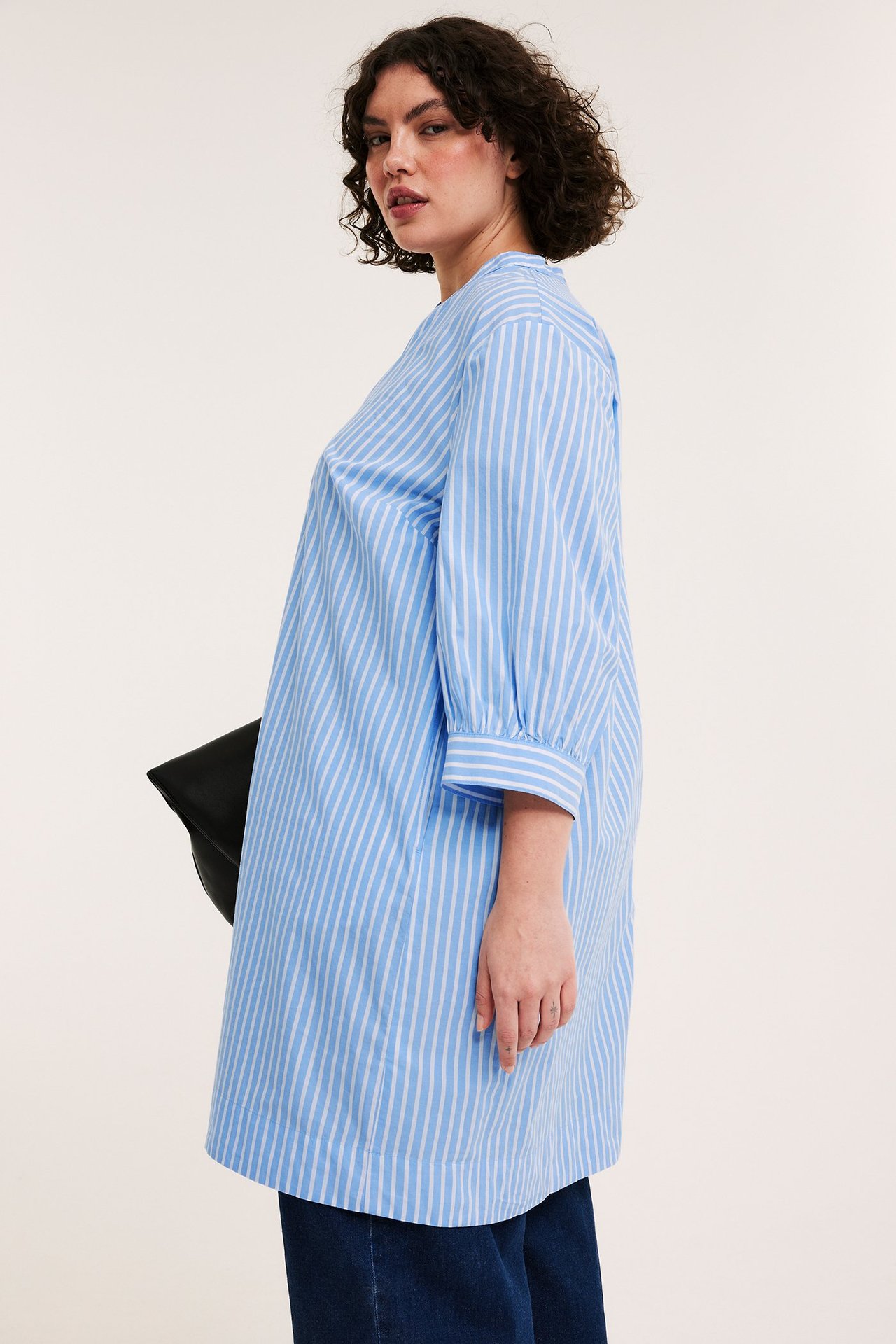 Sidovy av en kvinna i blåvitrandig bomullspoplinklänning med 3/4-ärmar, hållande en svart clutchväska.